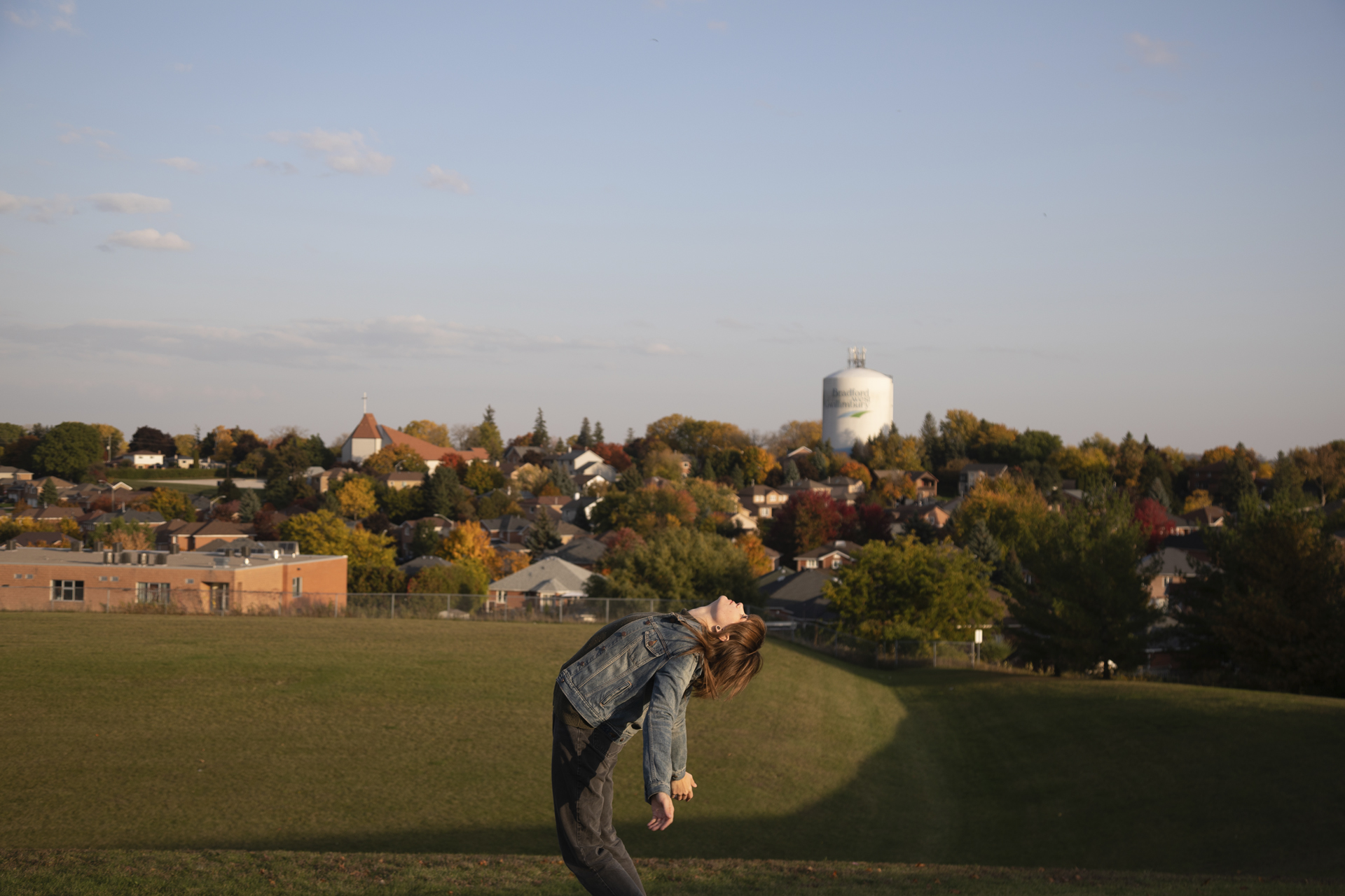 Over Backwards: a woman bending backwards on a hill with her arms dangling beside her on a sunny day. In the background there is a skyline filled with townhouses, trees, a church and a water tower.