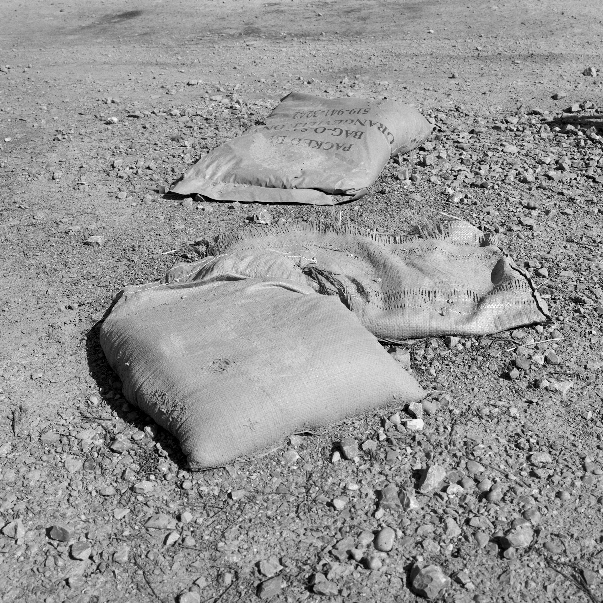 Where The River Runs # 2: A square black and white photograph of three bags of sand strewn about dirt and rocks. The material on the bags is fraying, and their contents are spilling out. A footprint from a construction boot can be seen on one bag of sand.