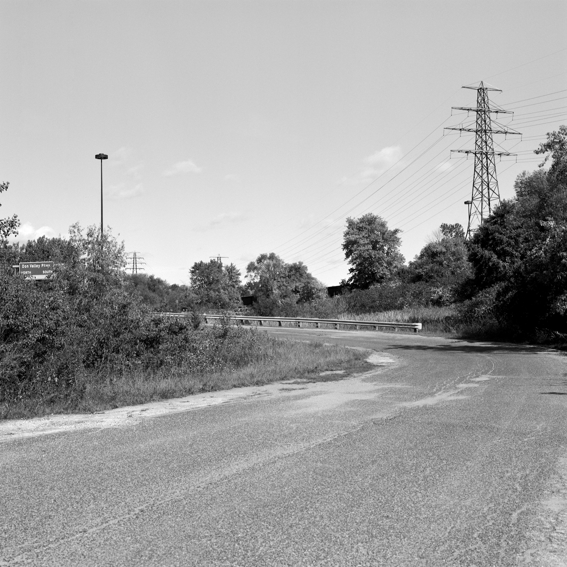Where The River Runs # 5: A square black and white photograph of a decommissioned roadway. The road extends from the bottom, leading into a guard rail positioned in the middle of the frame. A sign that reads 'Don Valley Pkwy' can be seen through the trees on the left side of the image as electric towers and power lines come from the right.