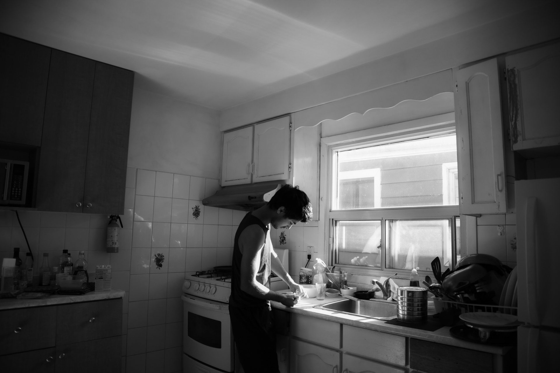 This photo consists of a man in a brightly lit kitchen preparing something to eat. He has short black hair and is wearing a sleeveless tank-top and pants. The photo is black and white.