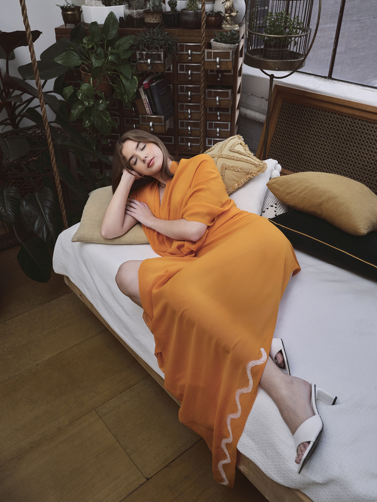 This image there is a Caucasian young woman laying down on a white sofa wearing an orange dress that flows off the bed. Behind her is a cupboard with plants and books. The Sofa is covered in pillows ranging in size and color between gold, green, and white. The young woman is laying with her eyes closed, presumably asleep