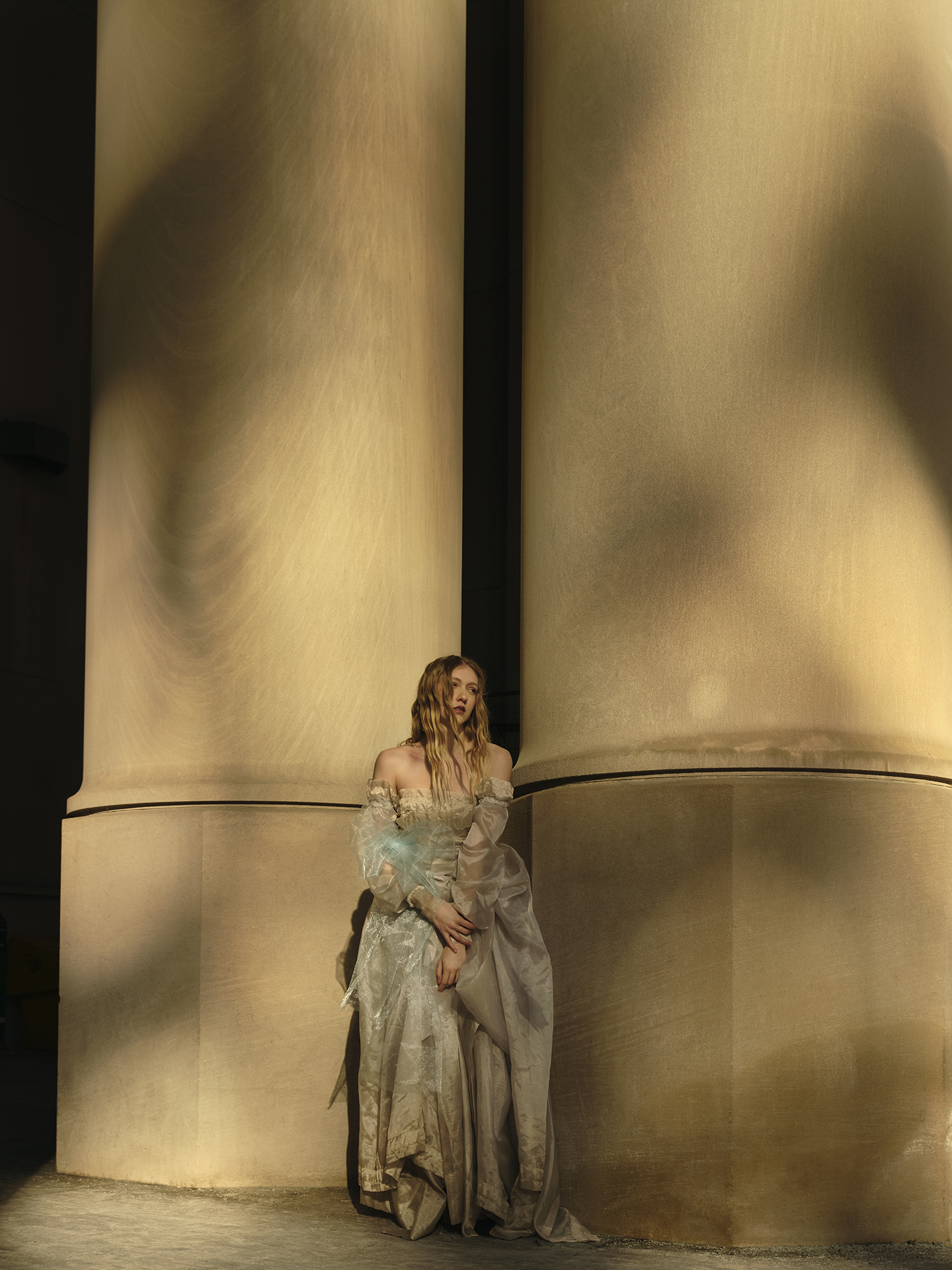 In this image, a Caucasian young woman is leaning against one of two pillars in the photo. The speckled creates a painterly effect on the pillars and adds separation between the subject and the background. She is wearing a seer and shiny dress with many layers, typical for the 18th century. Her blond hair softly blows in the wind as she gazes to the right.