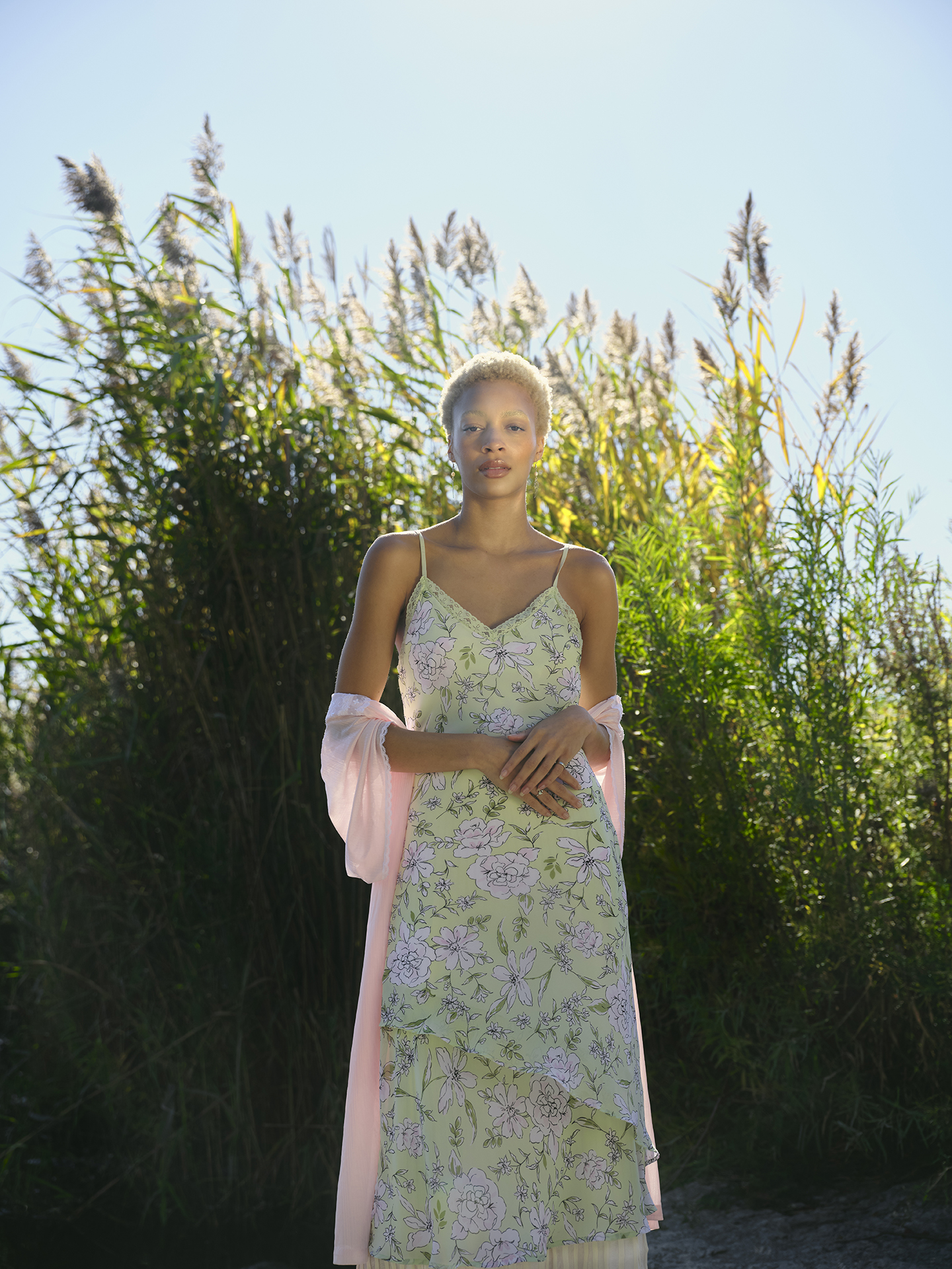 In this image, the subject is a light skinned model wearing a silky green dress covered in flowers with a pink scarf around her arms. She is standing in the middle of the composition facing the camera in front of lush green high grass that towers above her.