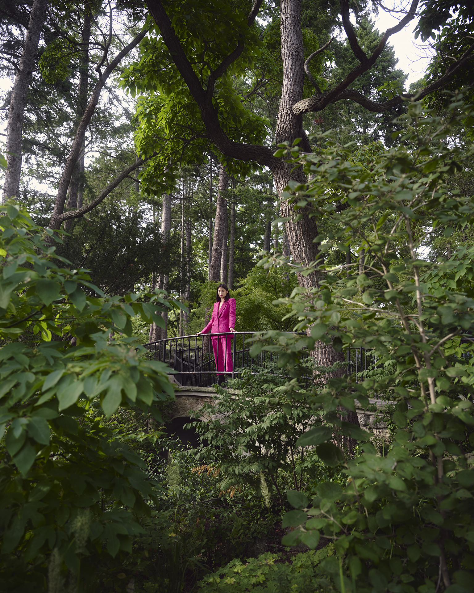 In this image, the female subject is wearing a pink suit in the center of the image. She is surrounded by foliage and greenery. The trees and plants are creating a spiral-esque shape that focusses into the female model.