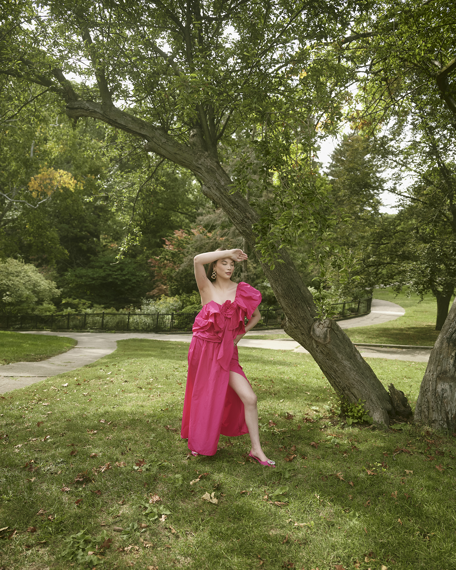 In this image, a female Asian subject is standing in the middle of the composition wearing a pink dress. She has one leg out of the dress’ slit and her right arm on her forehead. On her left side is a tree that grows diagonally across the image. Far behind her are three parkways and more greenery that take up most of the image.
