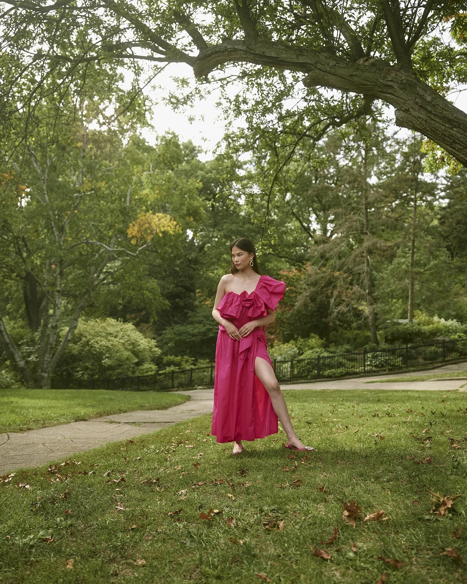 In this image, a female Asian subject is standing in the middle of the composition wearing a pink dress. She has one leg out of the dress’ slit and both her hands in front of her. On her left side is a tree that grows diagonally across the image. Far behind her are three parkways and more greenery that take up most of the image.