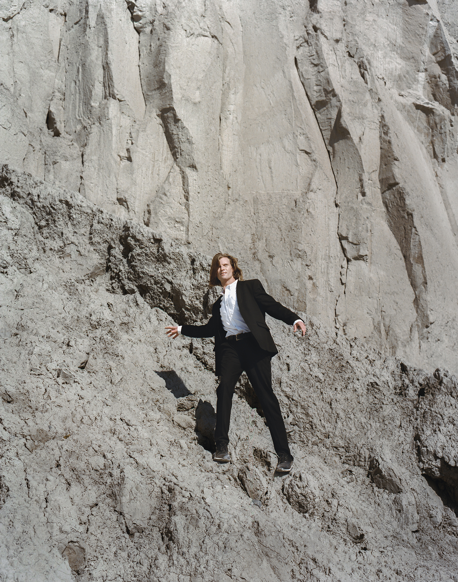 In this image, a white male can be seen wearing a black suit and white shirt on top of a cliff. His pose is awkward as he is trying to find balance as not to fall. He is surrounded with gray rocks that dominate the image.