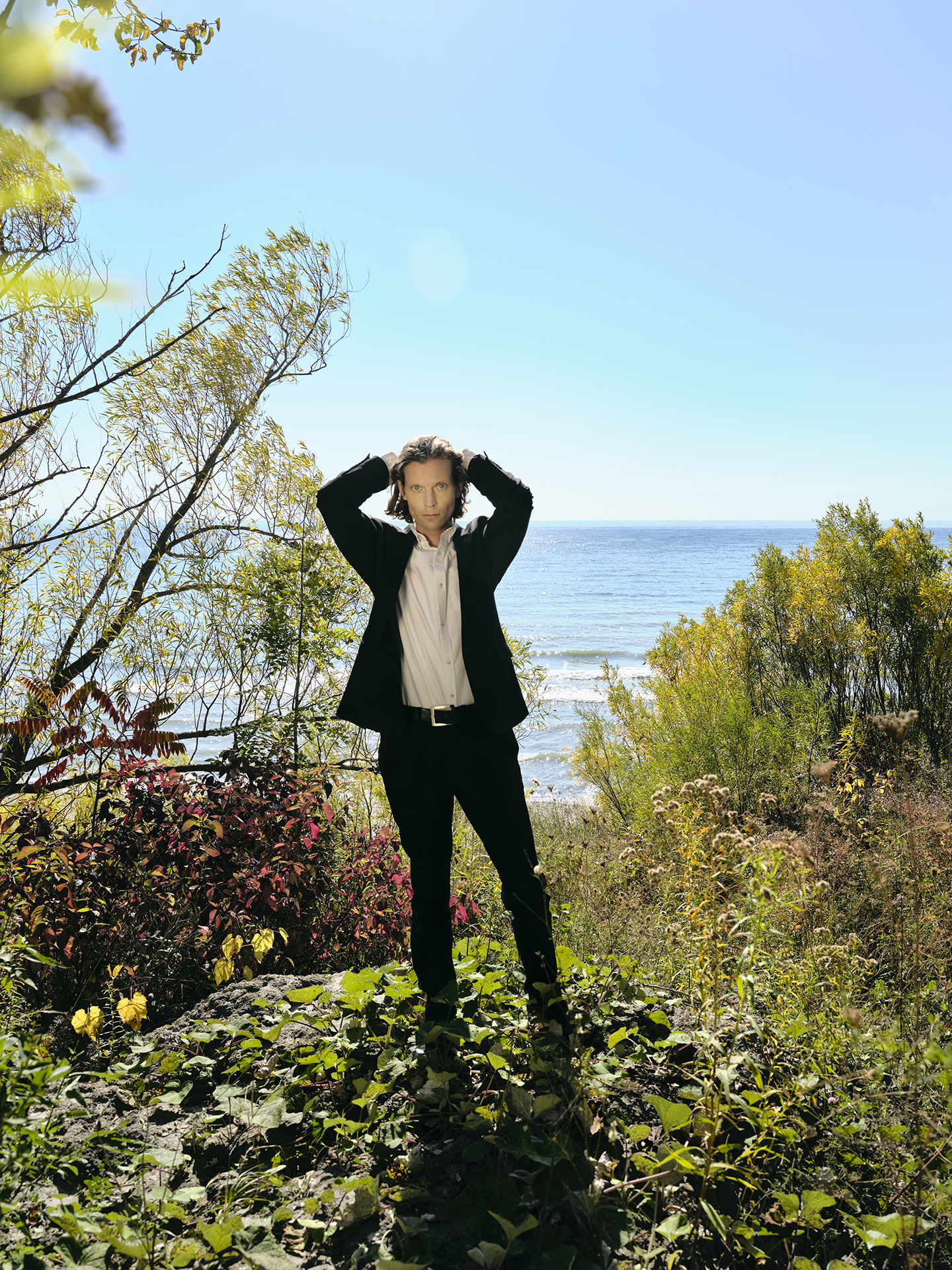 In this image, a male subject is standing centered in the composition wearing a black suit and white button up with both hands in his hair. He is standing on top of a cliff surrounded by greenery and plants. Behind him is a lake that goes to the horizon