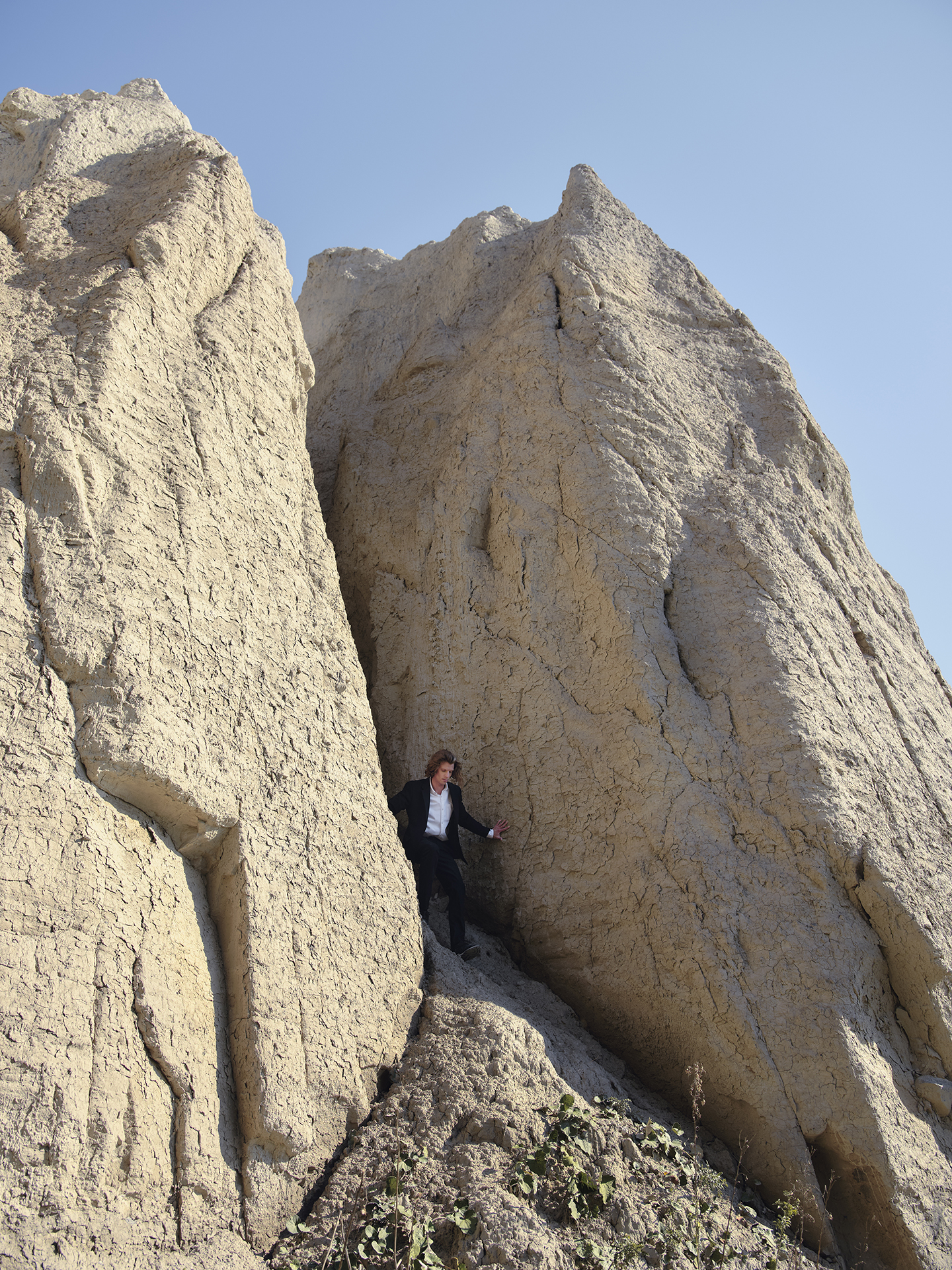 In this image, a white man wearing a black suit and white shirt is descending a spacing between two large stone structures. He is holding on to both sides and so he will not lose balance. The rocks have a grey- yellow tint to them that contrasts the blue sky.