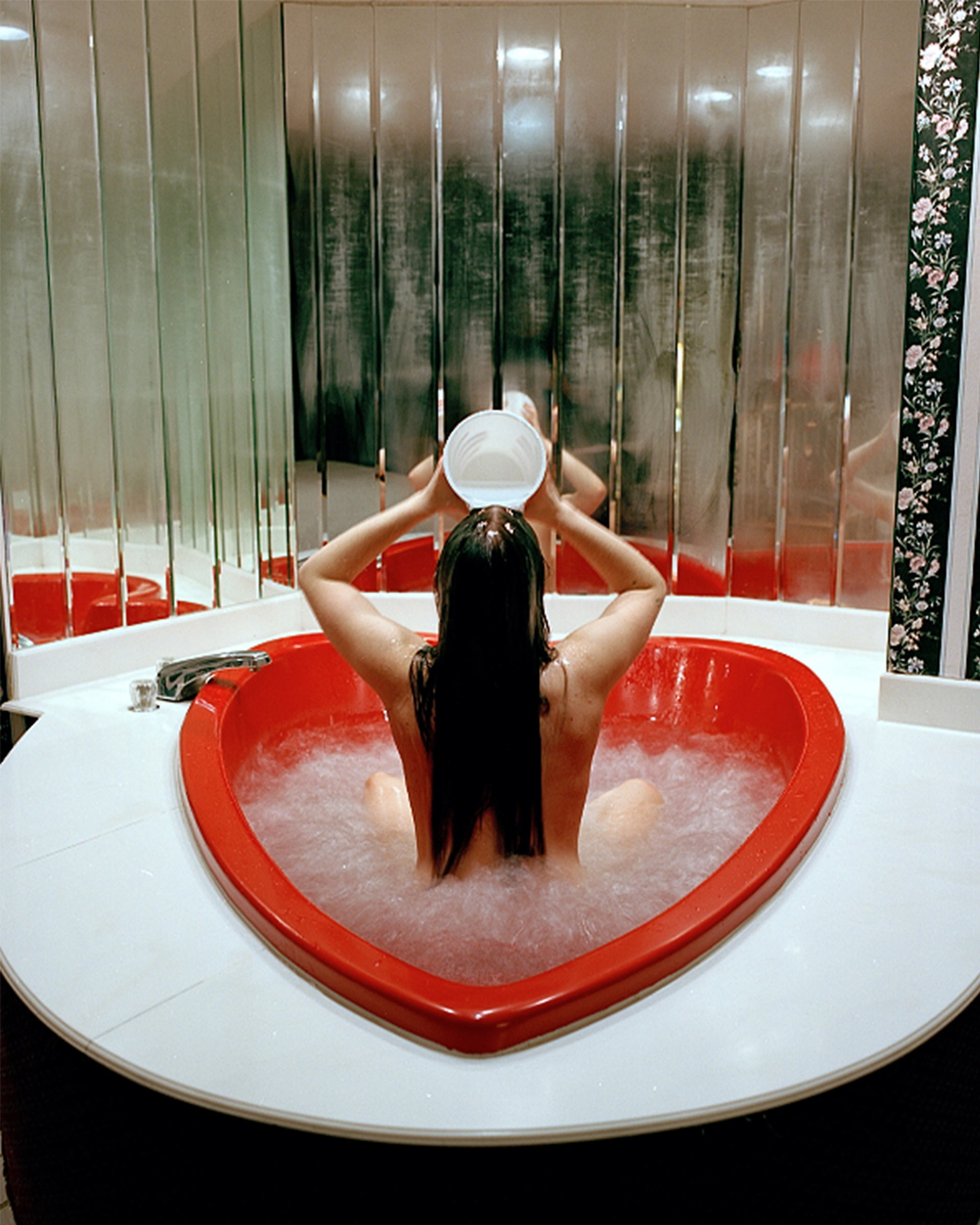 Love Tub: A woman facing away from the camera sitting in a hotel heart-shaped tub filled with water. She holds a white small bucket over her head and pours water onto her hair. A wall of fogged-up mirrors is behind her.