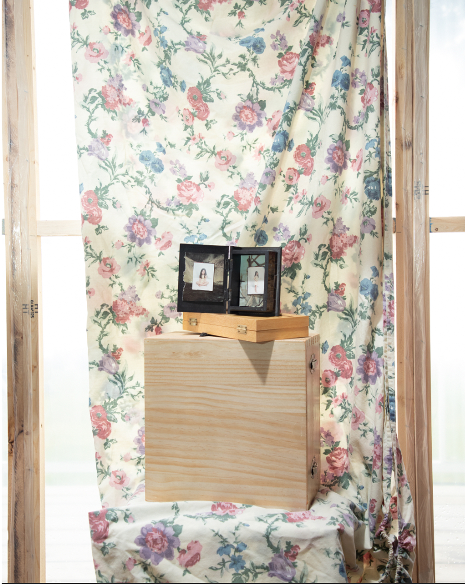 Untitled #1: A small box holds two archival family photos of a young girl. The box sits on a pile of wood in front of a floral sheet, inside a sunroom.