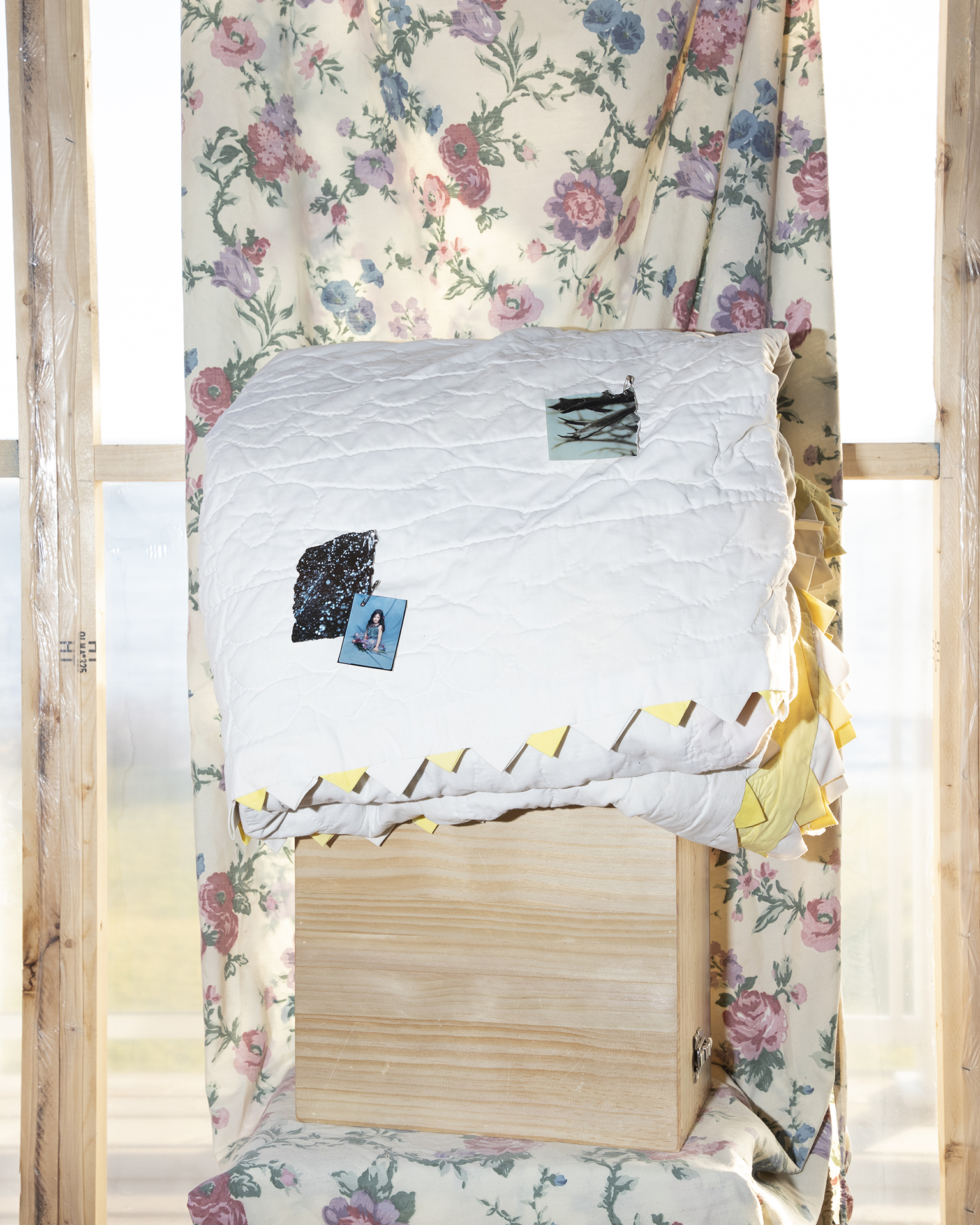 Untitled #2: A quilt with pinned-on archival family photos of a young girl. The quilt sits on a pile of wood in front of a floral sheet, inside a sunroom.