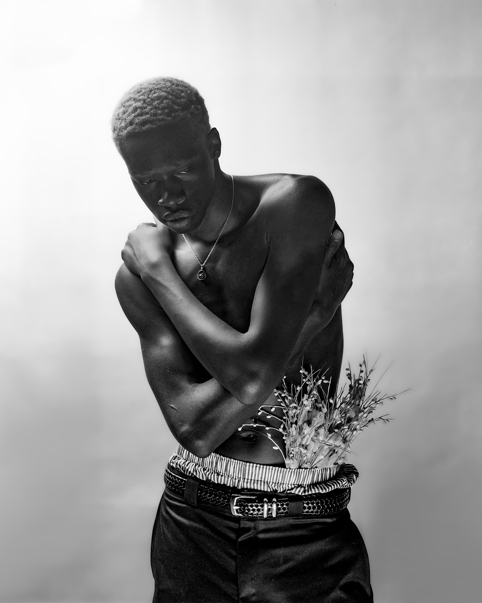 Flowers at the Door: Rukundo, a Black man with buzzed blonde hair, stands shirtless, with plants tucked into the waistband of his black pants. He is looking down, slightly hunched over while his hands are tucked on either side of his pants. He stands in front of a light-coloured background.