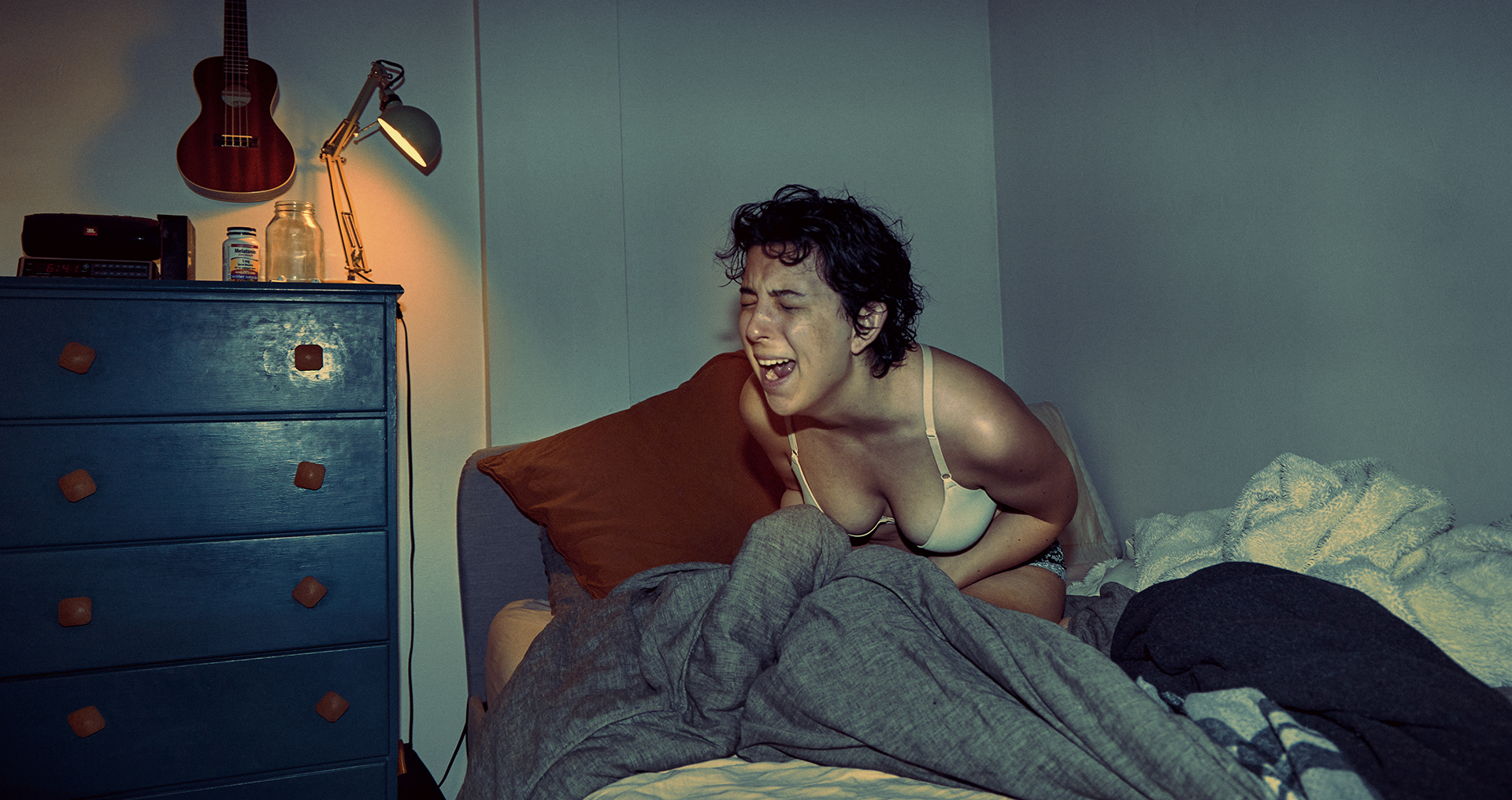 A photo of a woman sitting in her bedroom screaming out while clutching her blanket in her hands. The tone of the photograph is blue and there is a lamp emitting orange light on the left side of the photo.
