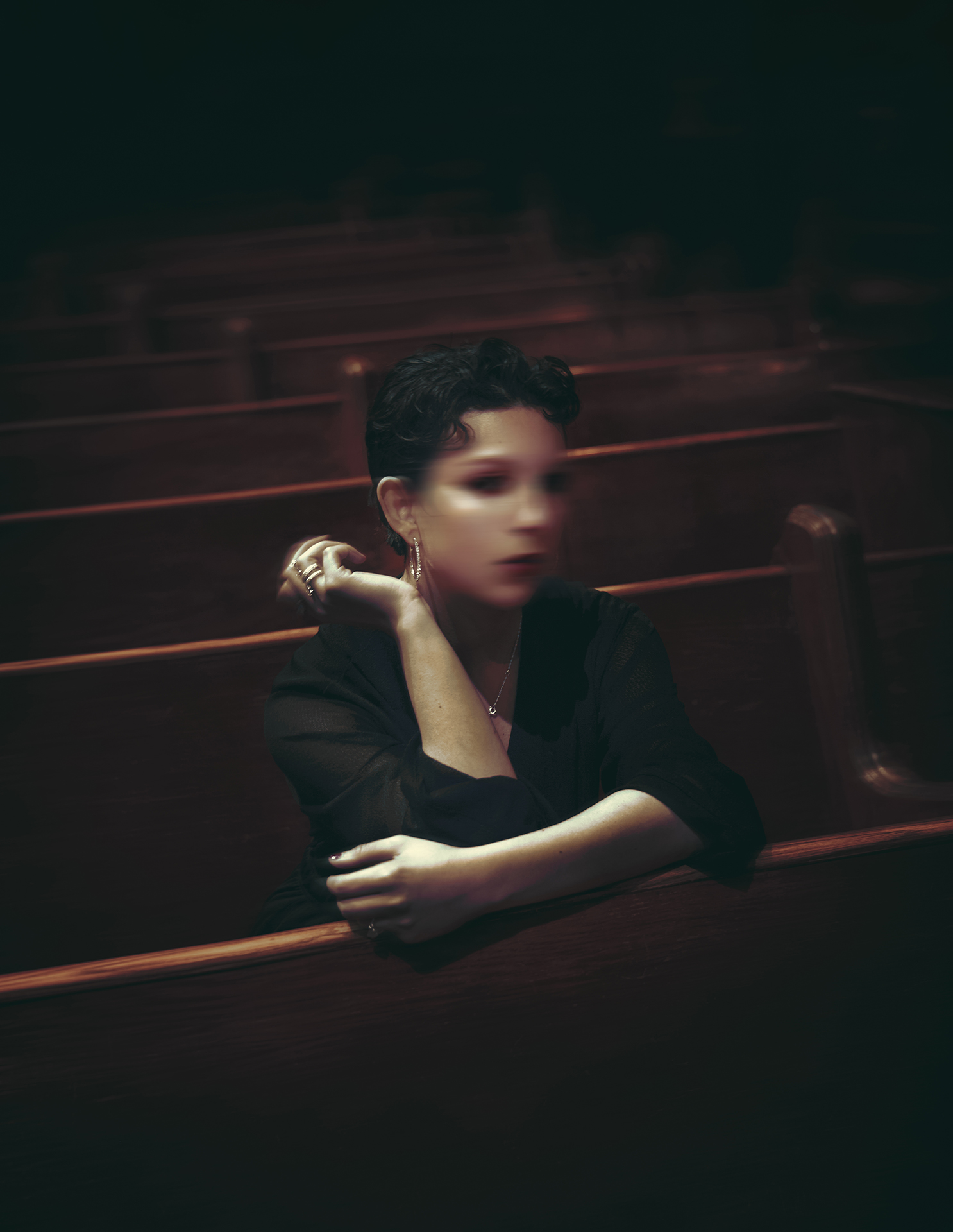 : A photo of a woman sitting in the benches at a church. She is wearing all black and her face is blurred out.