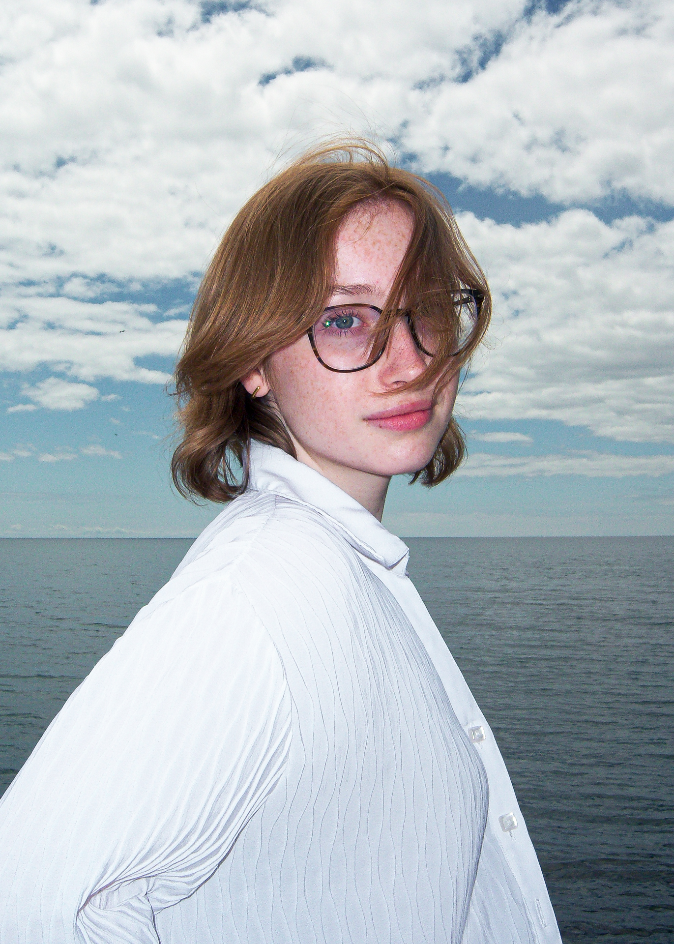 Flash Series The model, Regan, is standing on a beach wearing a white button up shirt and black rimmed glasses. She is looking straight forward as the wind blows hair into her face.