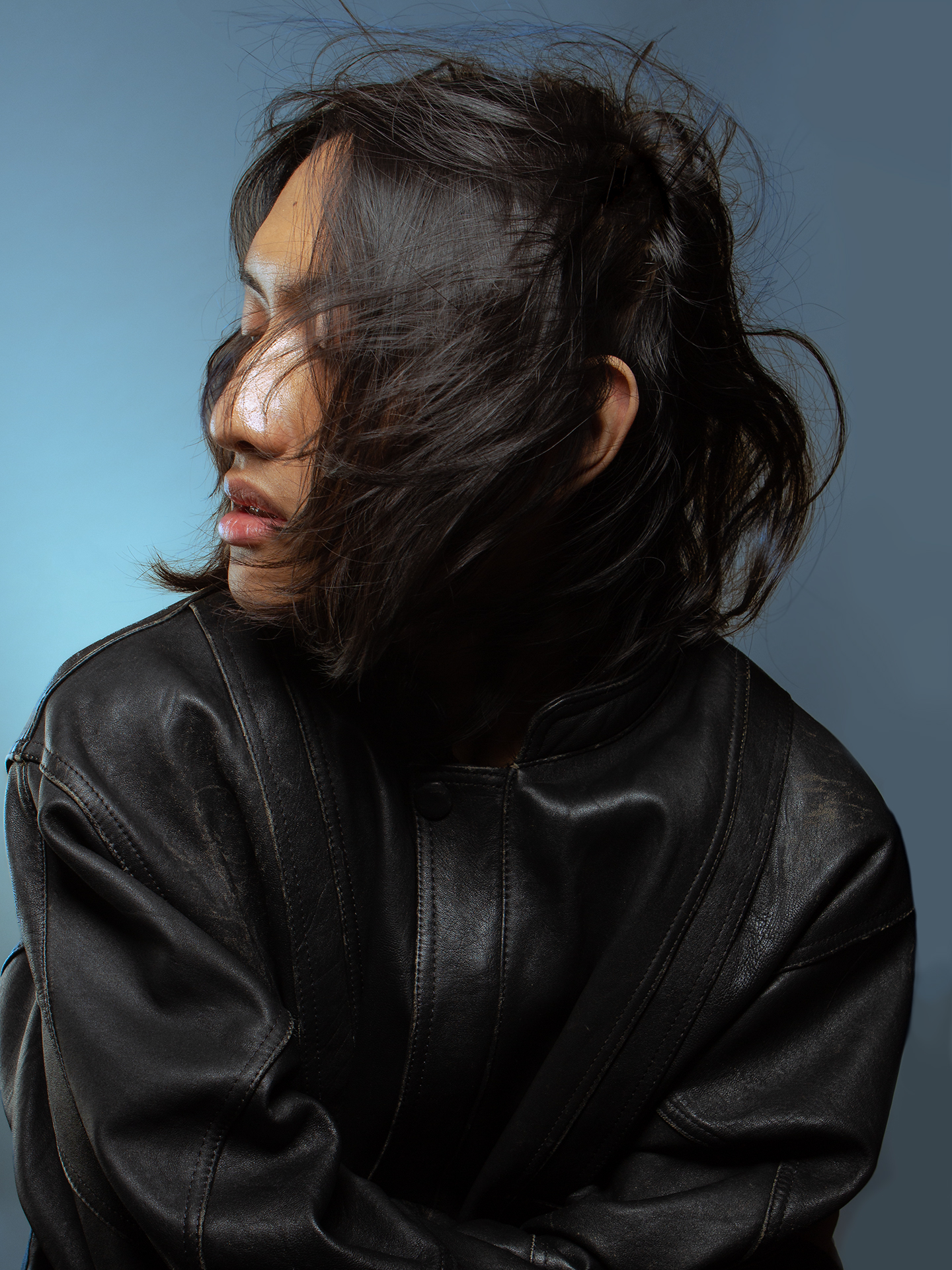 Sai The model, Sai, has their head turned to the left side of the image with their eyes closed, as their short black hair blows onto their face. Sai is styled in a black leather jacket against a blue backdrop.