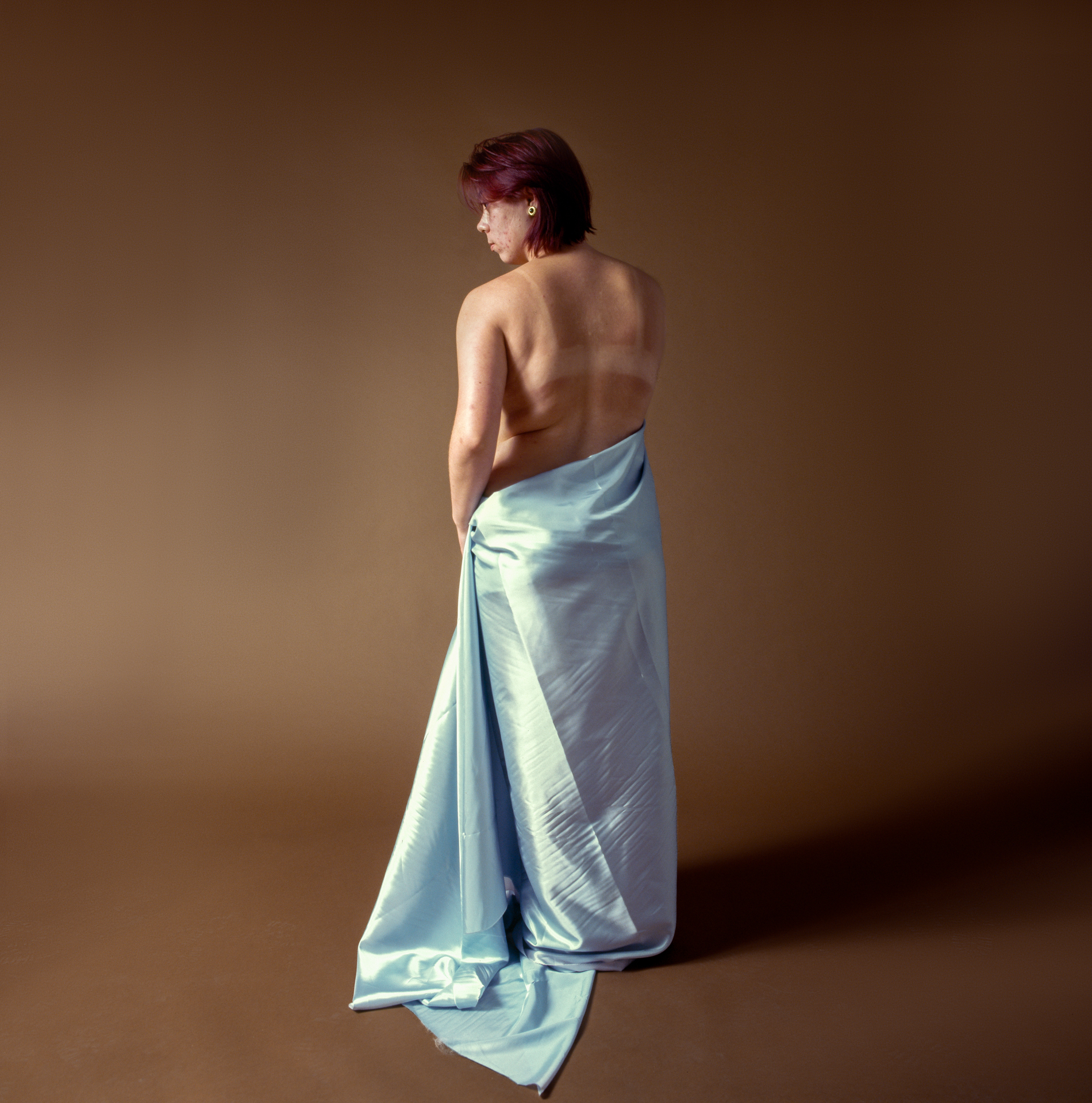 Cut From The Same Cloth An image of a young woman with short red hair stands with her sunburnt back towards the camera, the lower half of her body only covered by a blue sheet.