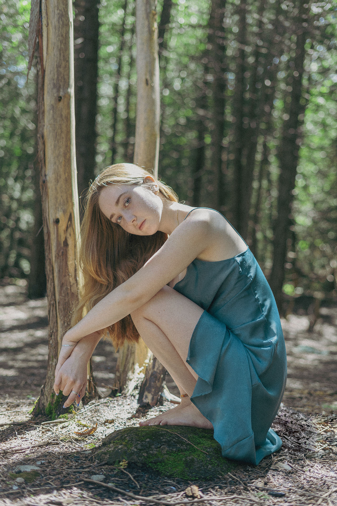 A young woman in a forest in a teal dress crouches on a mossy rock while bare foot