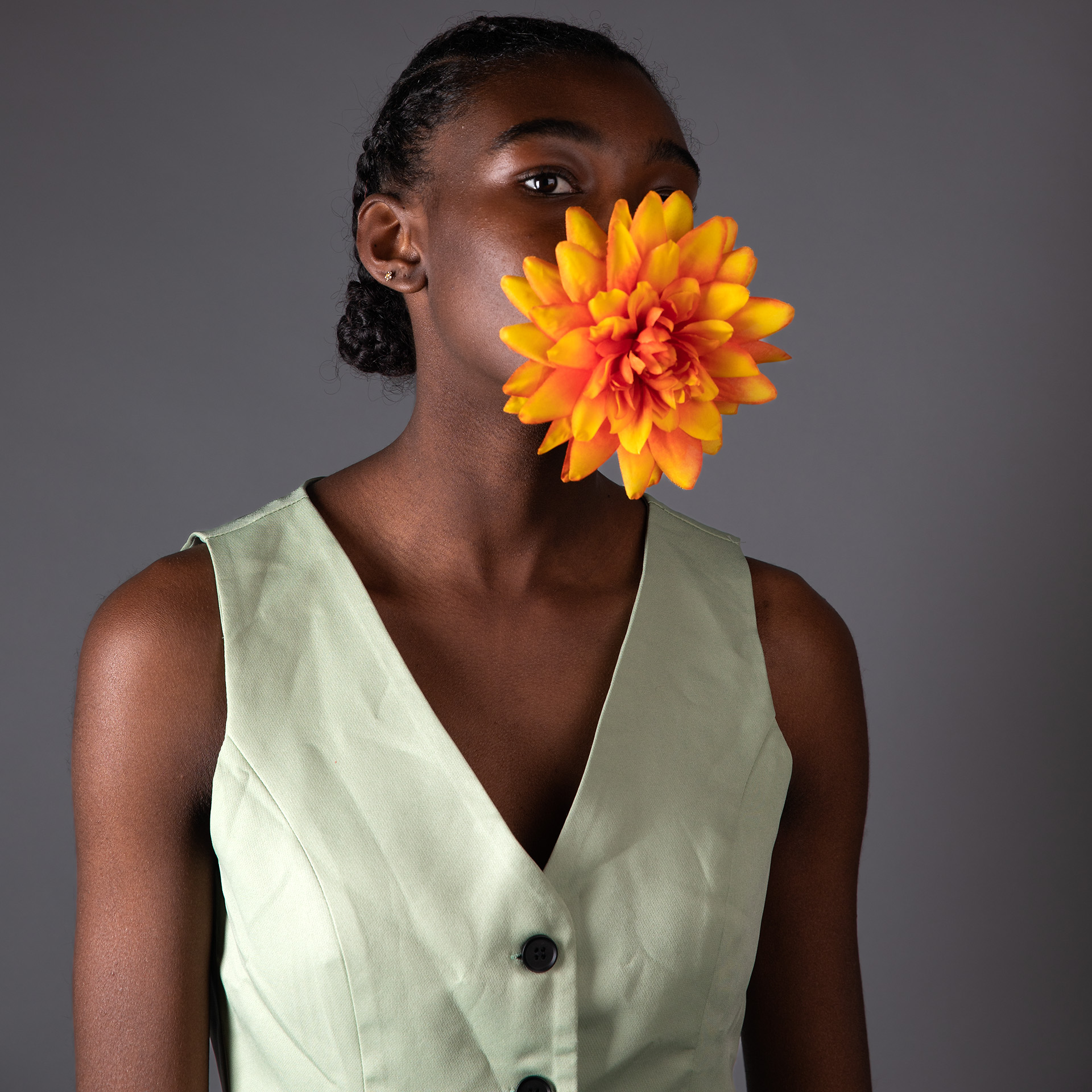 Black woman, her hair styled into short braids that are wrapped in a bun. She is wearing a light green vest 1 and a half buttons are in frame. One eye is looking directly into the camera, the other eye is blocked by a petal of an orange flower in her mouth.