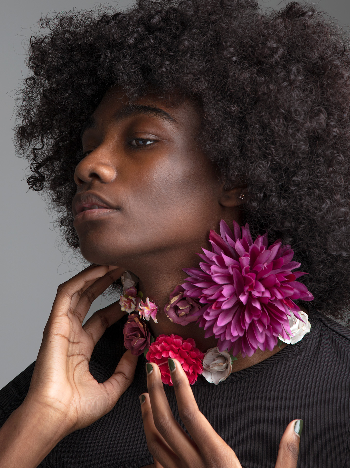 Black woman, with her hair styled in an afro, her left hand is softly touching the side of her neck, her right hand is in front of her. Her gaze is cast downwards. She is wearing a black shirt. Eight flowers of different sizes and species grow on her neck in colours ranging from pink, white, burgundy and purple.