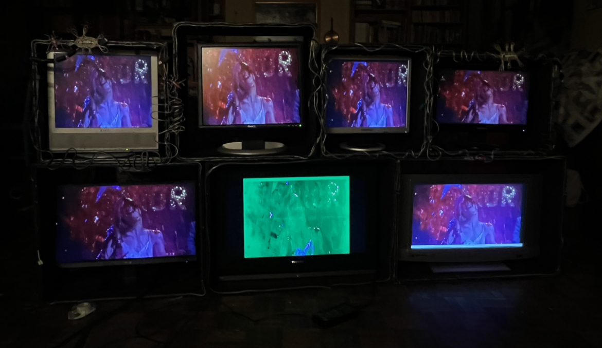 This image shows the structure which holds 7 monitors of all different sizes, decorated with scrap wires and the previously mentioned neurones. The monitors are displaying a still visual from Carrie 1978. This image is in darker lighting to better show the visuals on the screen.