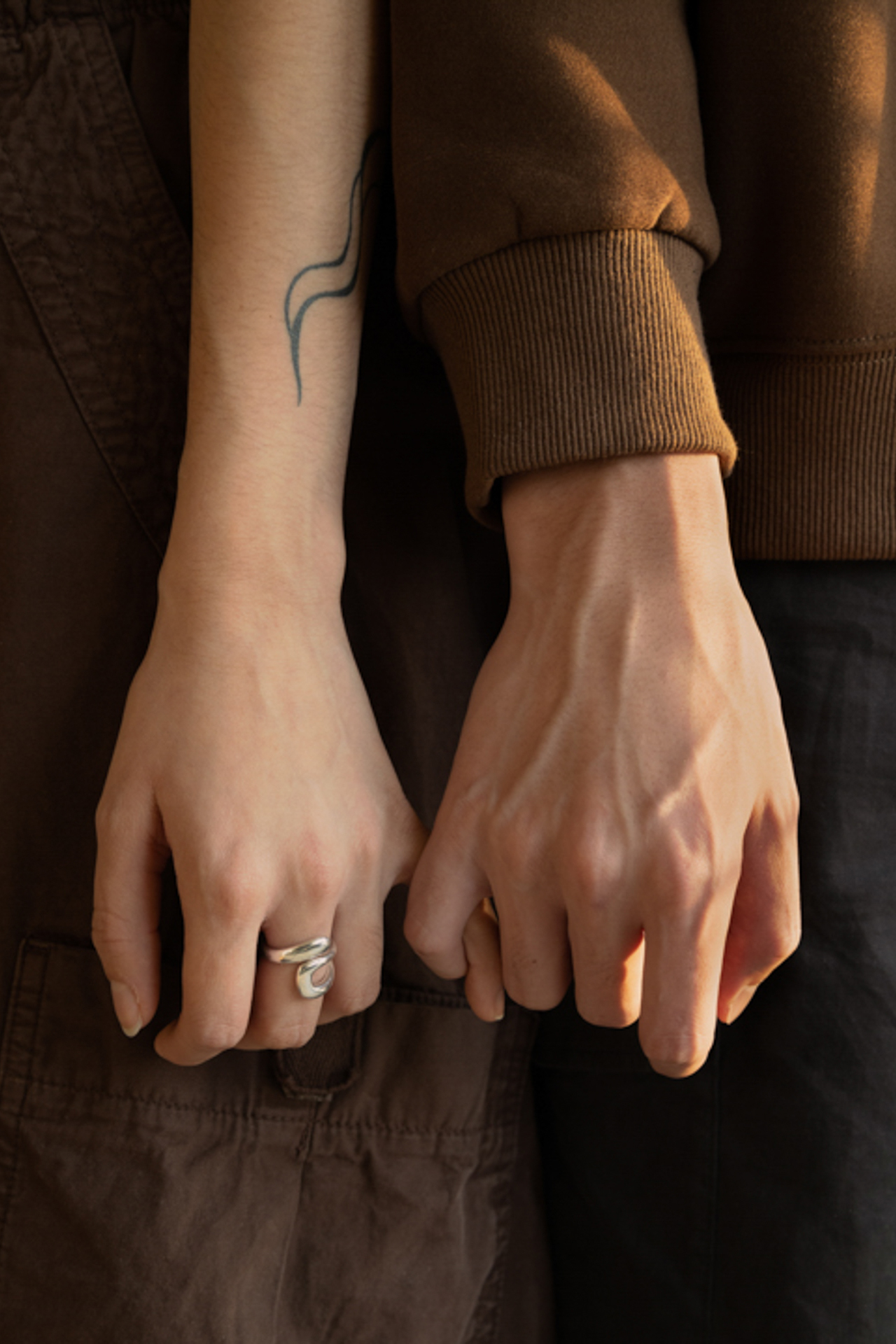 this photograph is a close up detail shot of two people's hands linked by their pinkies. The hand to the left is wearing a silver ring on their middle finger. The hands are at waist level and the overall colour scheme consists of warm browns and greys.