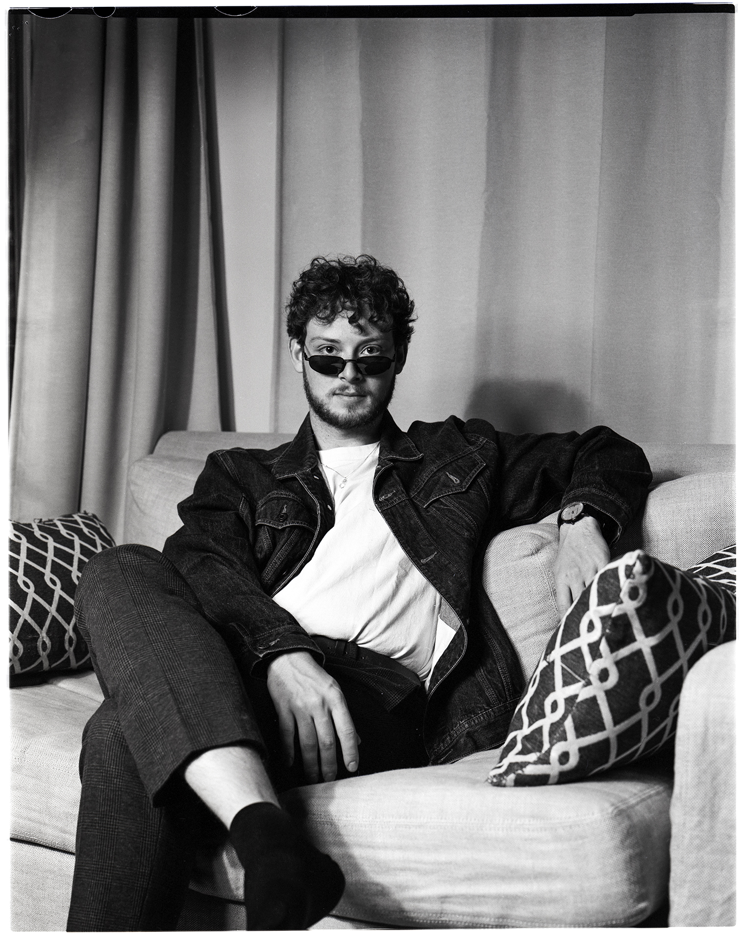 : A black and white image shot on film depicting Jamie, a man in his early 20’s with a beard and short curly hair. Jamie is sitting on a couch in front of a curtain looking directly into the camera. His legs are crossed and he has a straight face. He is wearing a denim jacket with a white t-shirt underneath paired with dark pants as well as thin sunglasses under his eyes.