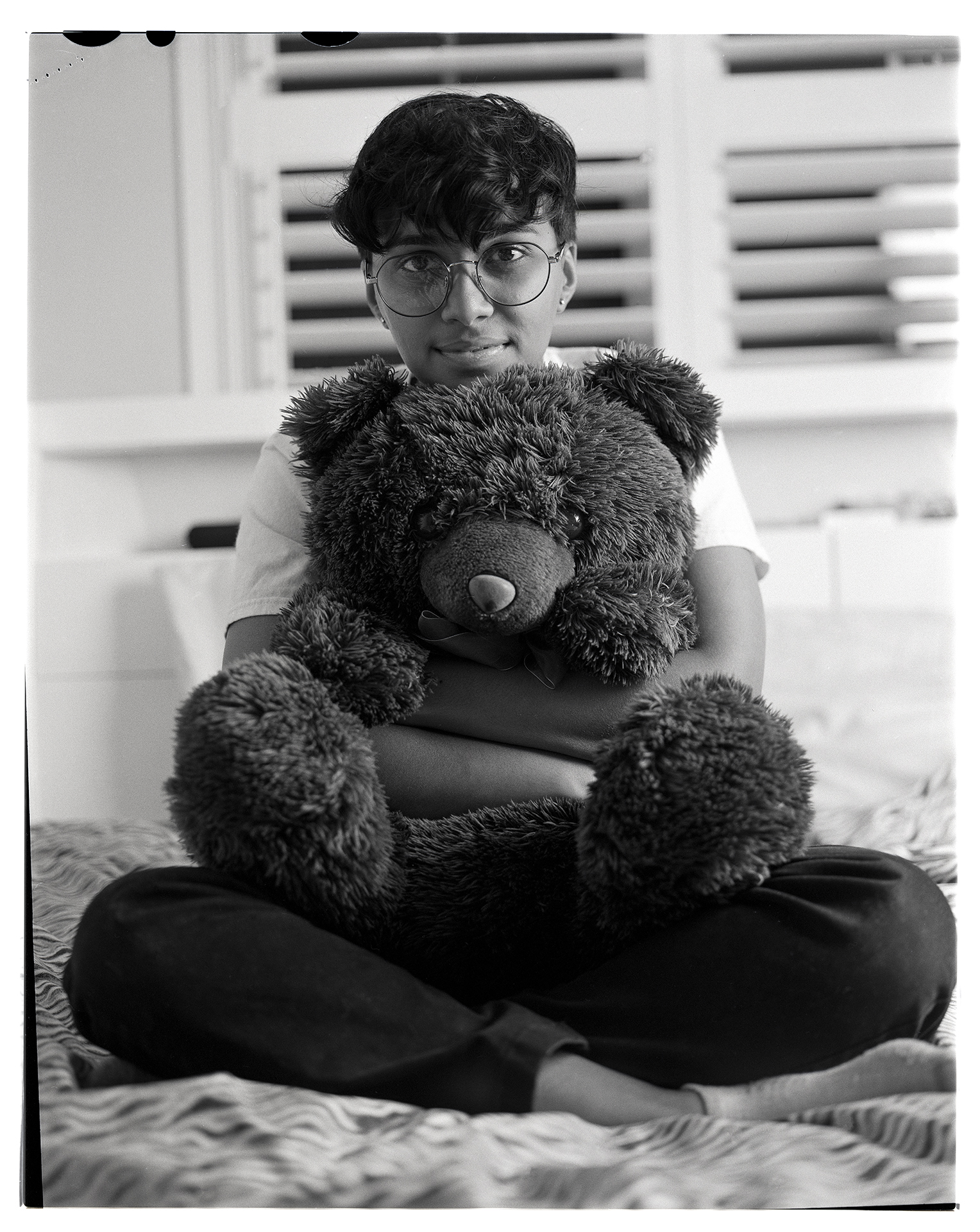 A black and white image of Shikha, a woman in her early 20’s with short wavy hair. Shikha is sitting on top of her bed holding a large dark teddy bear. Her head is resting on top of the bear while her arms hug it from behind. She is wearing dark pants and a white t-shirt.
