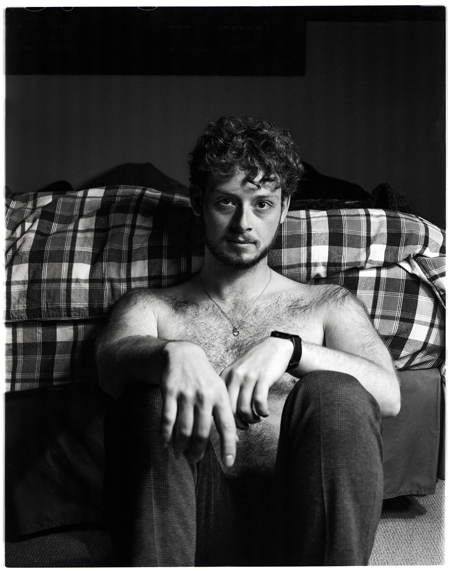 A black and white image shot on film depicting Jamie, a man in his early 20’s with a beard and short curly hair. Jamie is sitting shirtless on his bedroom floor wearing dark pants, directly in front of his bed. He is sitting with his knees together and his arms crossed while looking into the camera.