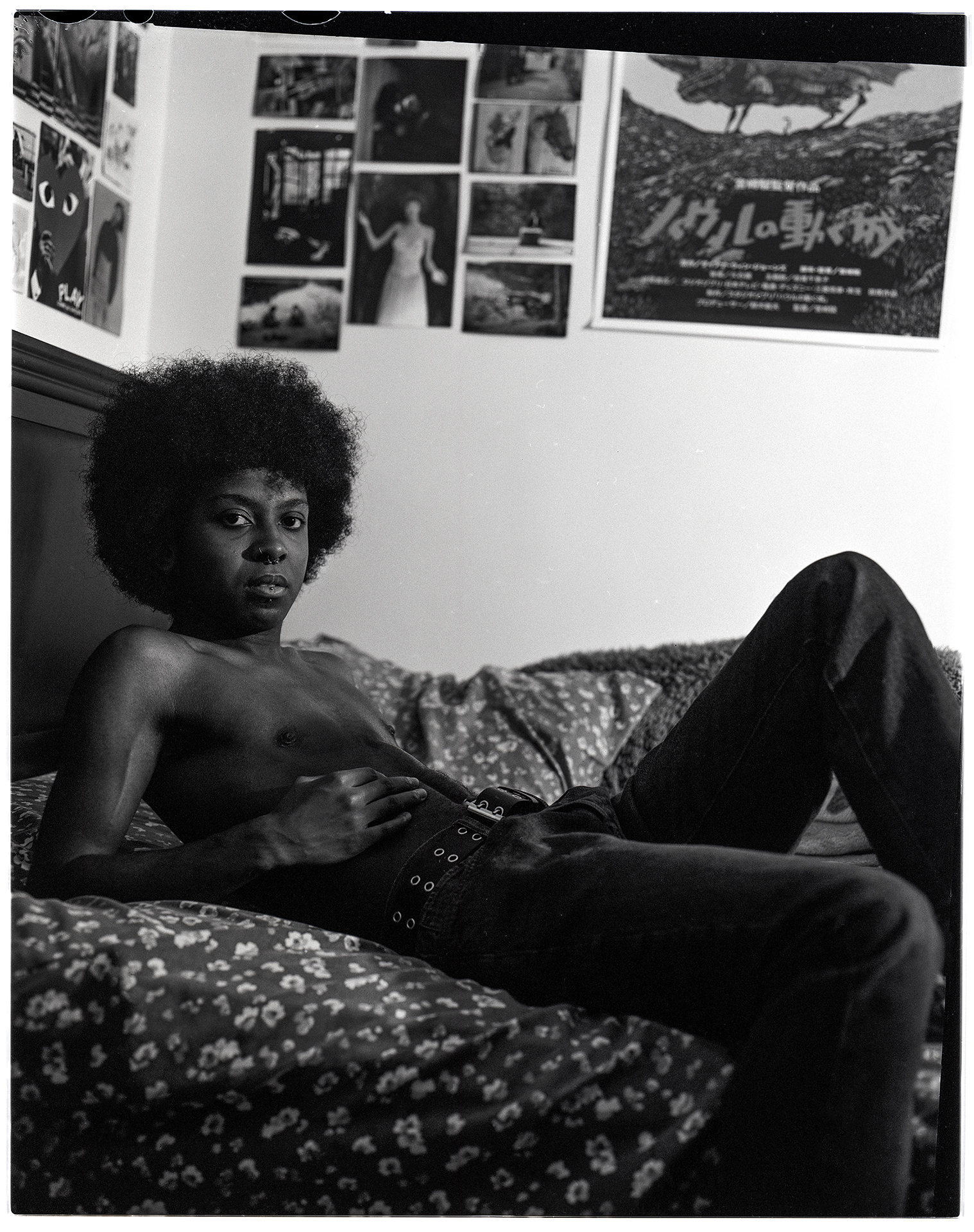 : A black and white image shot on film depicting Jayden, a person in their early 20’s with an afro. Jayden is laying shirtless on their bed. They are leaning back on their elbows while looking directly into the camera. They are wearing dark jeans and a belt.