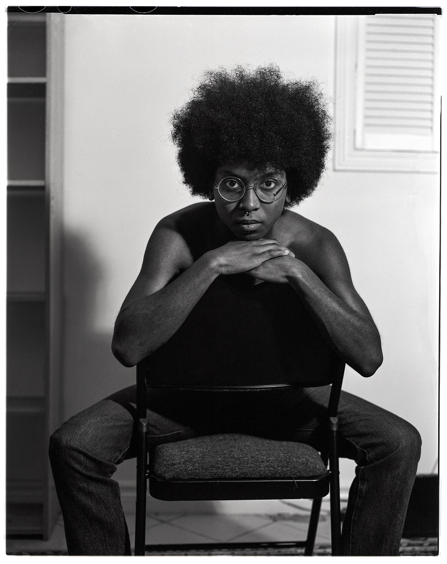 : A black and white image shot on film depicting Jayden, a person in their early 20’s with an afro. Jayden is laying shirtless on their bed. They are leaning back on their elbows while looking directly into the camera. They are wearing dark jeans and a belt.