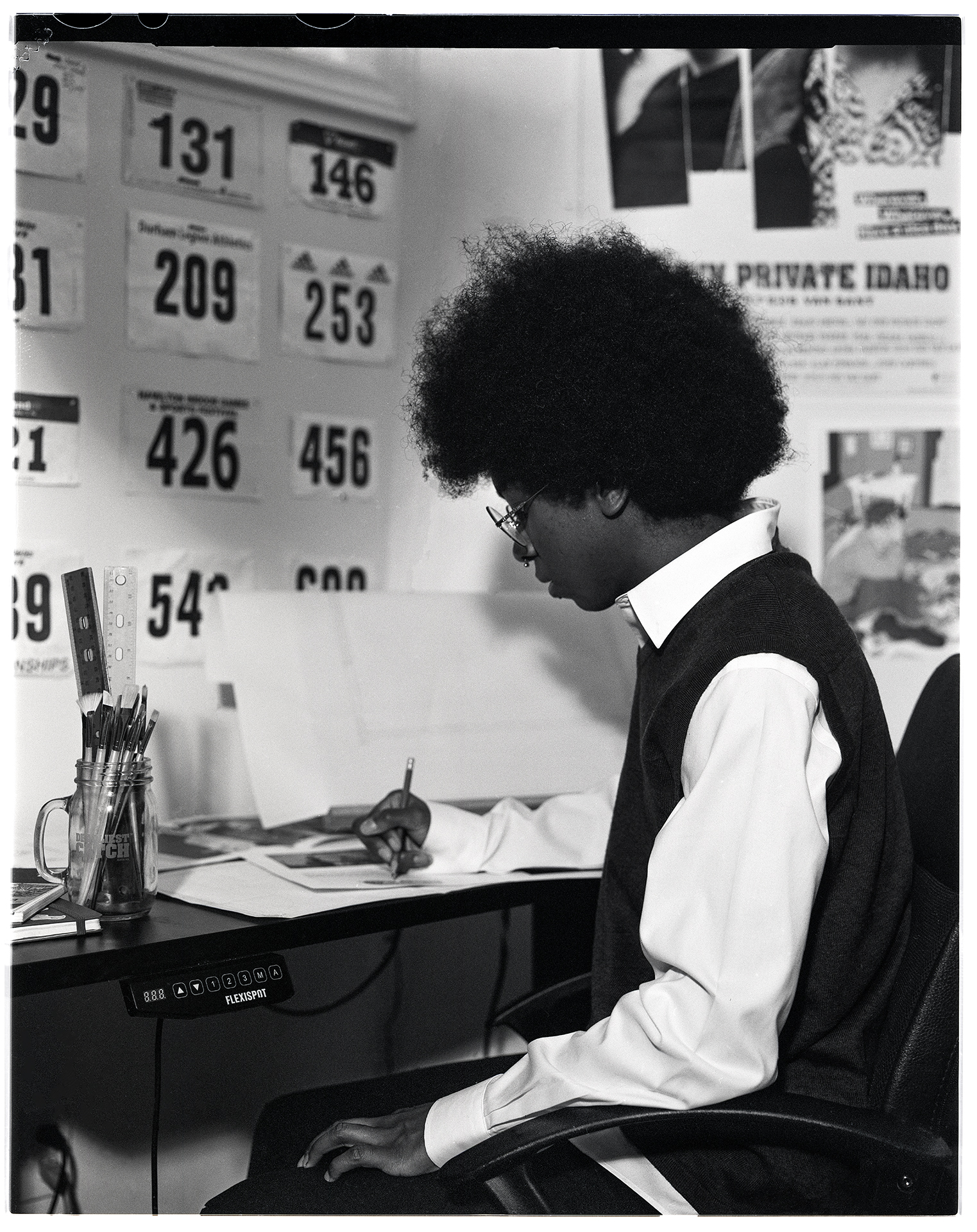 A black and white image shot on film depicting Jayden, a person in their early 20’s with an afro. Jayden is sitting at their desk. They are leaning overtop of a piece of paper while drawing. They are wearing a dark sweater vest with a white button-up shirt underneath.