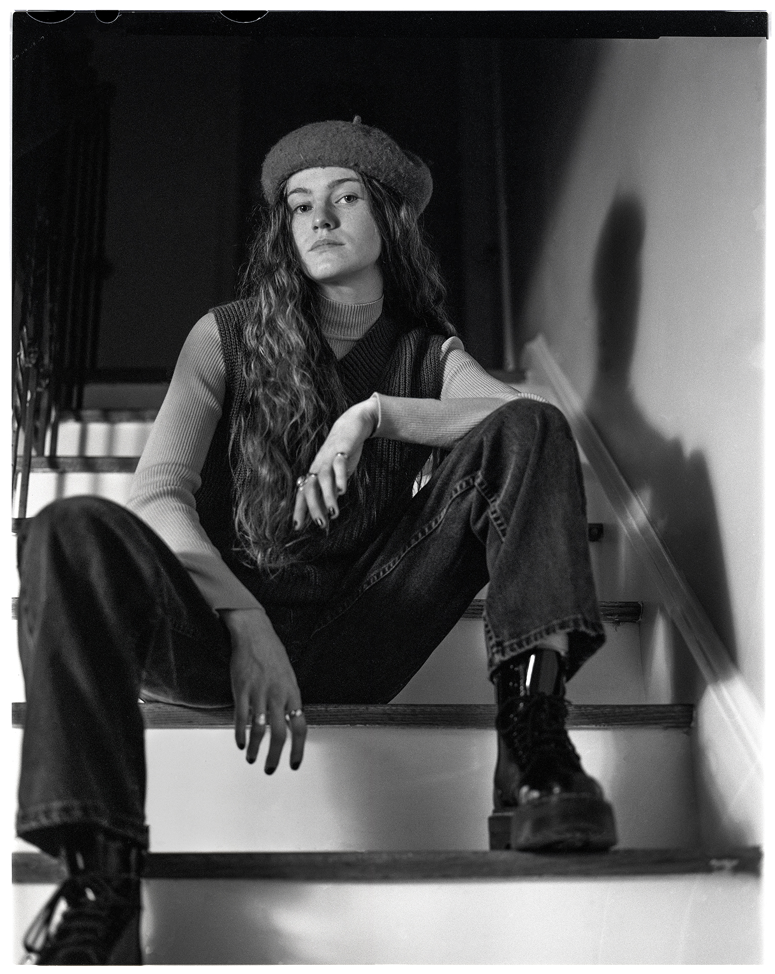A black and white image shot on film depicting Sam, a person in their early 20’s with long wavy hair. Sam is sitting on a staircase. They are leaning forward slightly above the camera with one leg positioned higher than the other. Their arms are resting on each leg. Sam is looking directly into the camera. They are wearing Doc Martin boots and a beret hat with a dark sweater vest and a light long-sleeve shirt underneath.