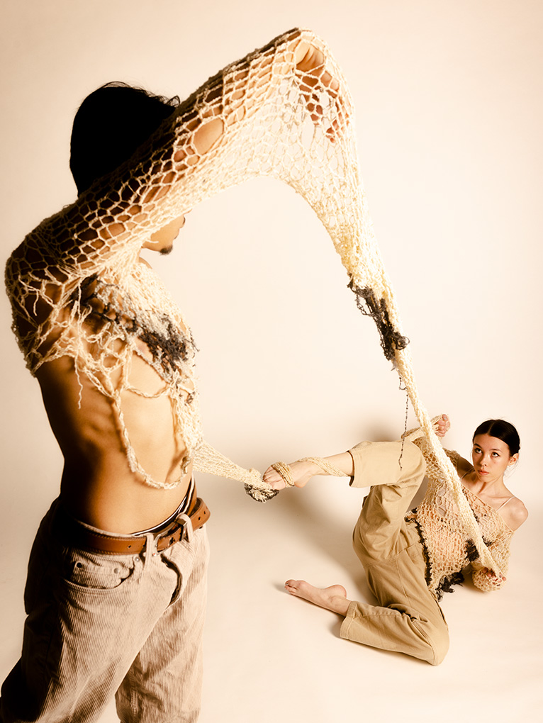 A photograph of two people intertwined by a garment piece. The sleeve of the garment is connected. The first model is standing and on the left side of the frame and is closer to the camera and the second model is sitting on the floor further from the camera.