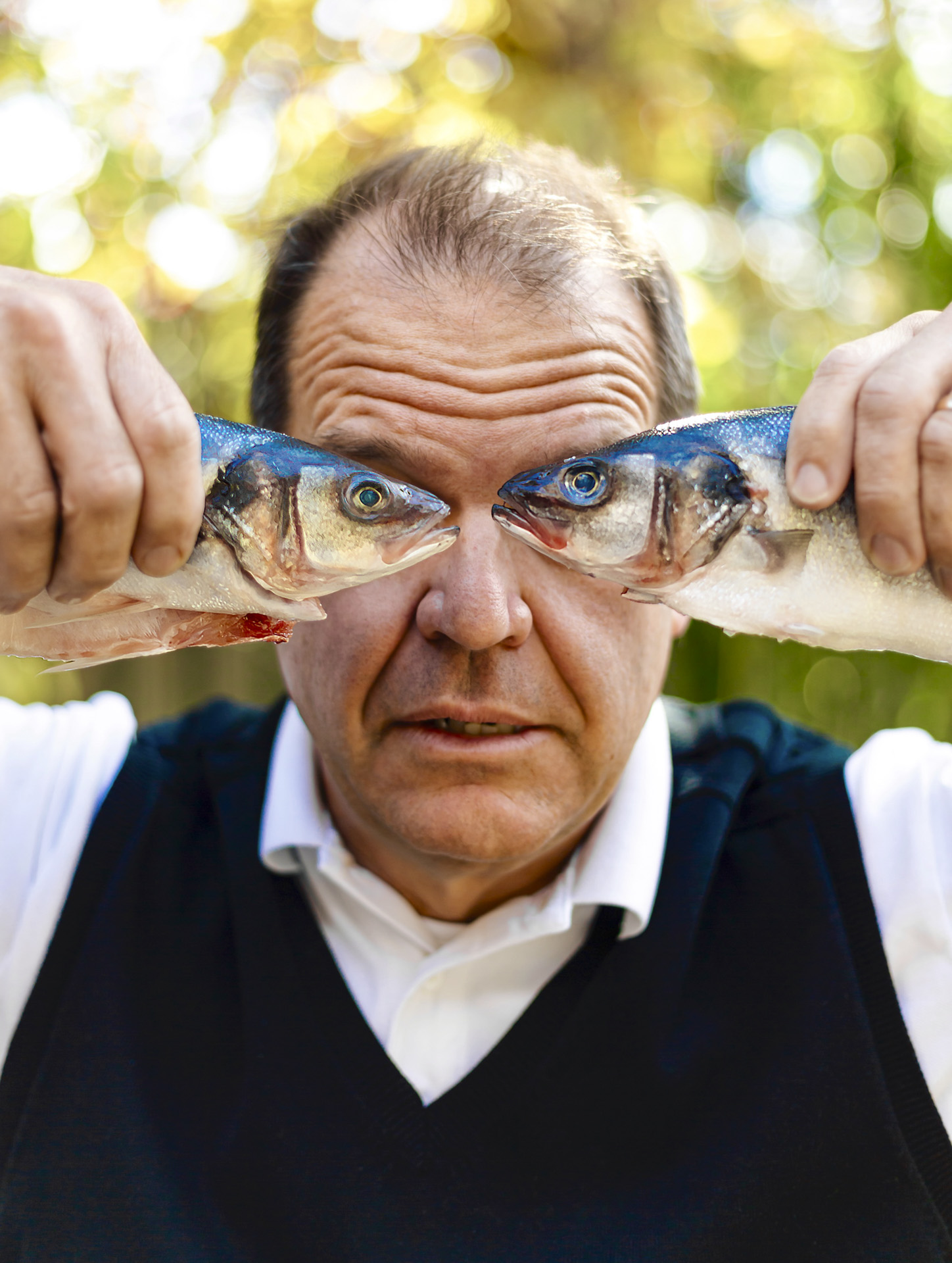 Fish Eyes: A middle-aged, medium-light skinned man is holding a dead fish up to his eyes in each hand. The man is wearing a white collared shirt with a navy blue sweater vest on top, looking directly at the camera.