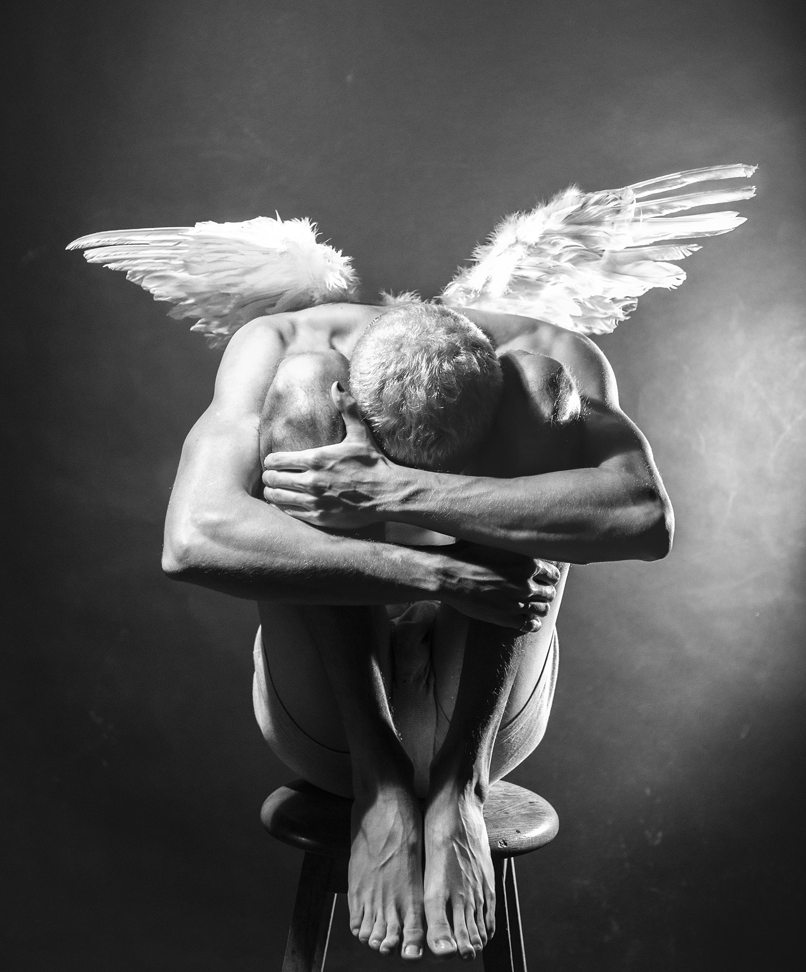 Mémoire Poétique A masculine figure is huddled on a bench in a position of self-soothing. A pair of wings stick out from his back, glowing with light.