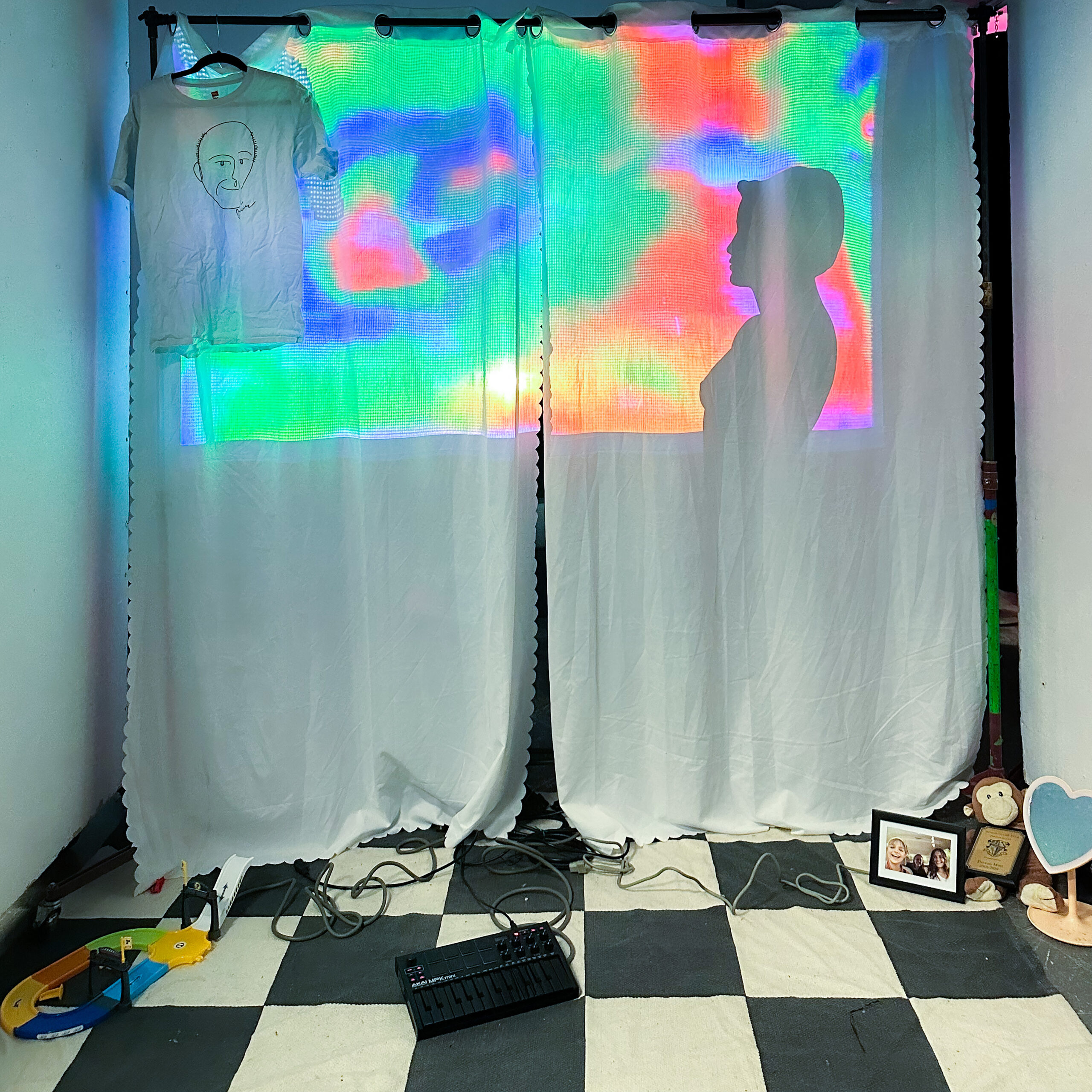 interactive installation with a projection through white curtains. there is a checkered rug with personal items on top (monkey plush, framed photo, heart shaped mirror, midi keyboard, kids toys, exposed wires)