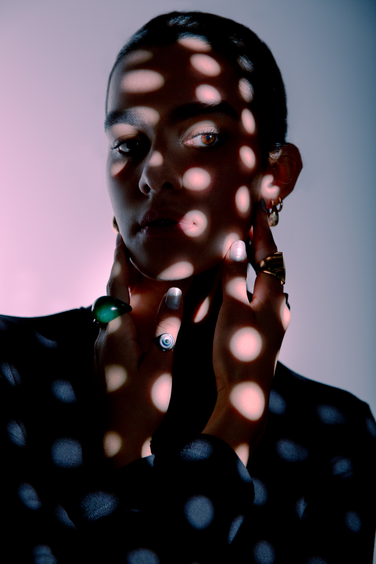 Adriana is posing with a circular pattern of lights hitting her face and upper body. She's wearing a black full sleeve top. Her accessories and eyes shine through in the light against a gradient background.