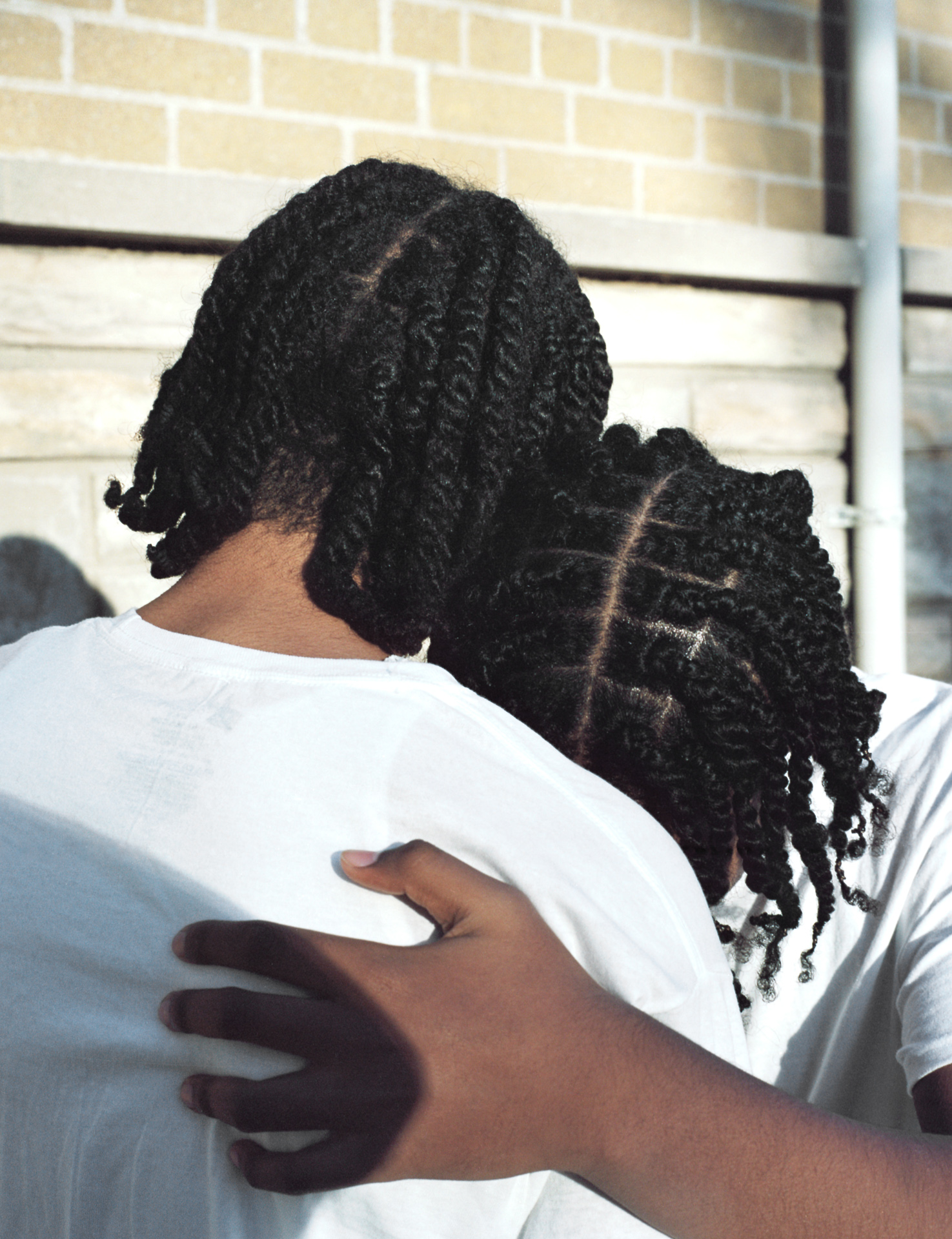 Acceptance: Nathan and Jare, both Black men with mid-length twisted hair are outside on a bright day. They wear white short-sleeved t-shirts and hug each other, facing away from the camera. Jare's hand on Nathan's back and the shadow of Rahim's elbow as well.
