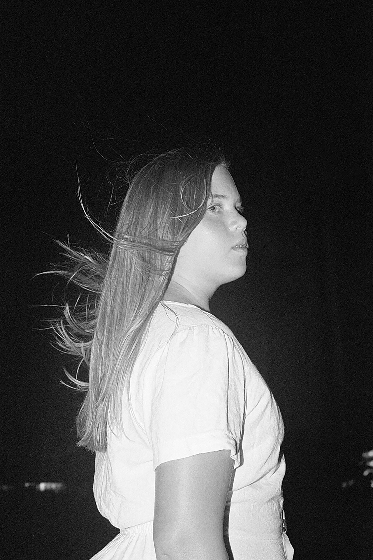 A black and white image of Eden in the middle of the frame, surrounded by a dark sky. Her hair is blowing in the wind, and she gazes off past the camera lens.