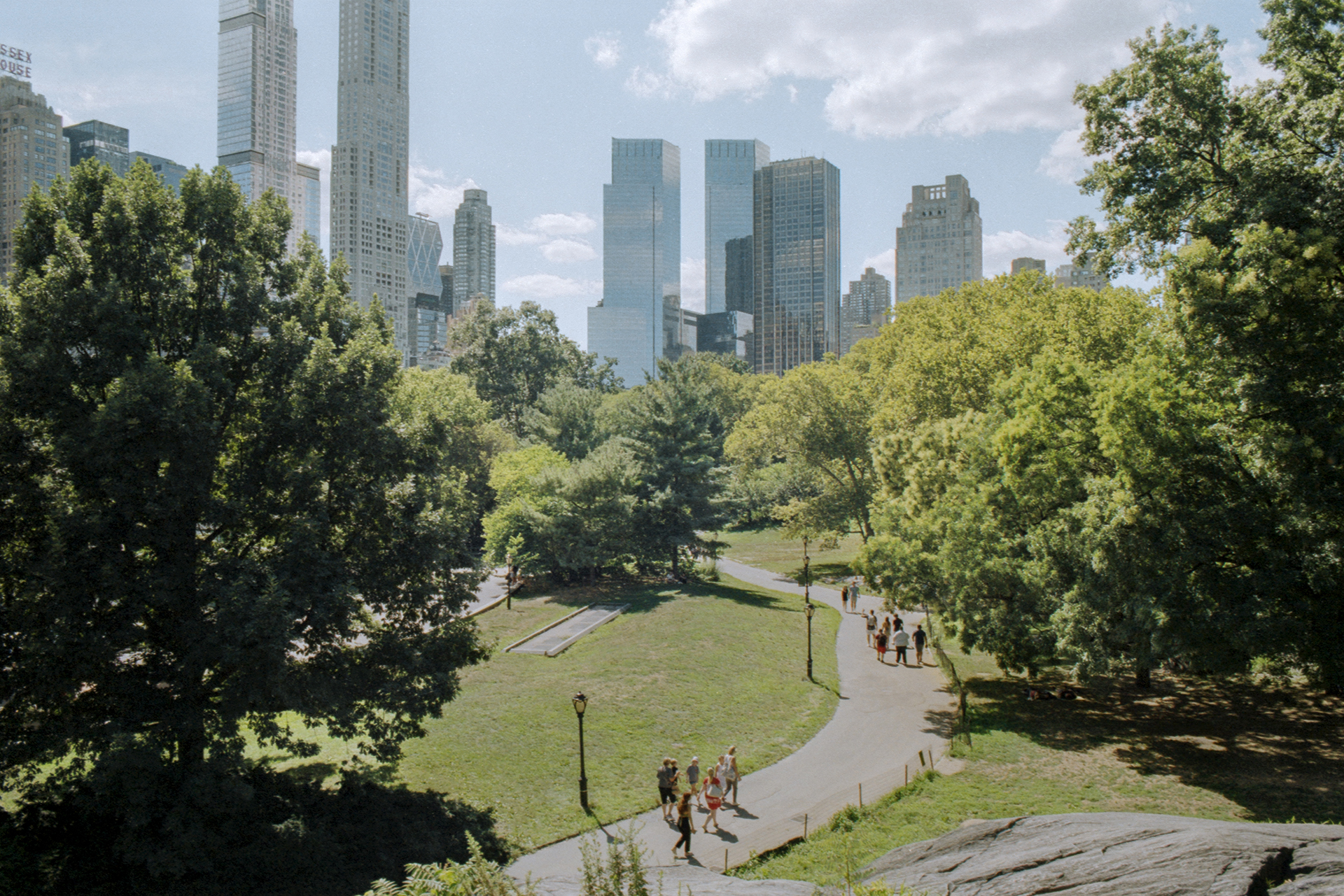 A colour image of a pathway in Central Park New York. On the S-shaped path are two groups of people walking down with a significant space between them. In the background the the Skyscrapers tower above the greenery and plants of Central Park