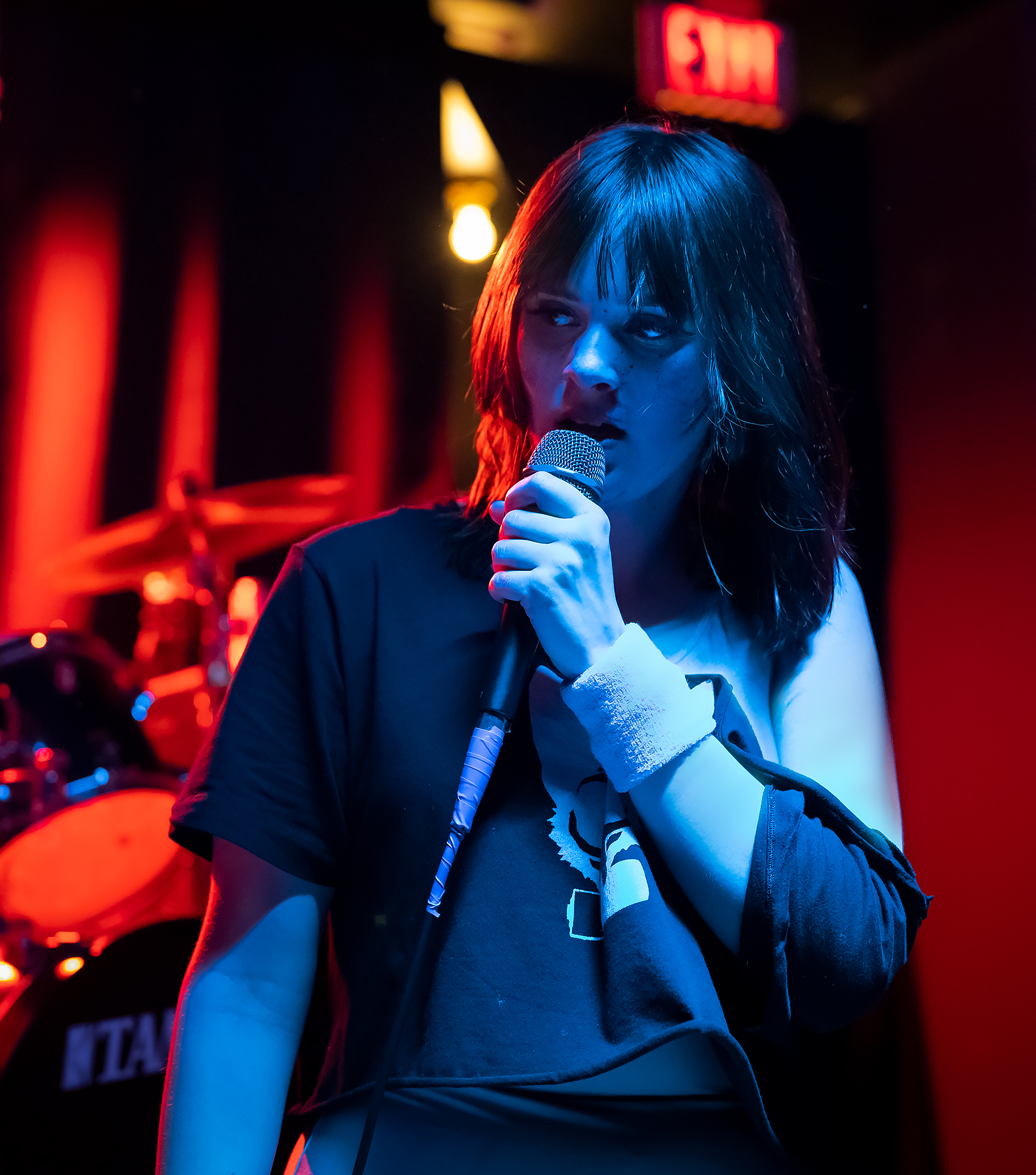 Redd, lead vocalist for band "Electrokat", is posed singing mid-performance wearing 80s style workout gear. She looks off in thought in front of a drumset, featured in contrasting red and blue lights.