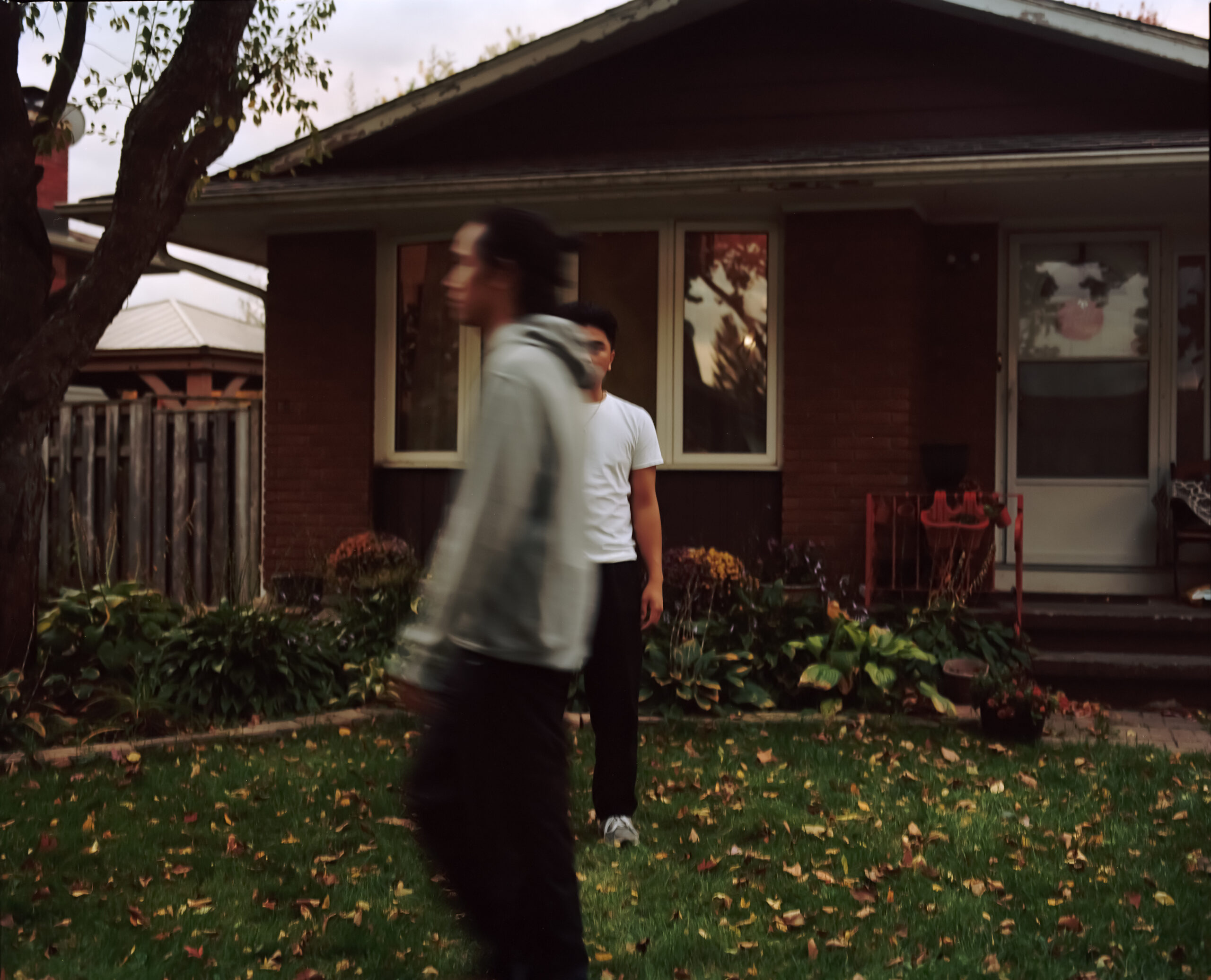 In front of a house A blurred person walks by a man looking into the camera