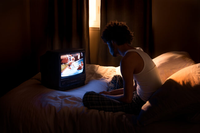 A man in a white tank top sits cross legged on top of a bed. Golden window light seeps through the closed curtains behind him. His face is illuminated by the blue light of a CRT television. Pictured on the television is a scene of a young man jumping into a pool.
