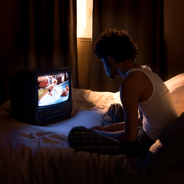 A man in a white tank top sits cross legged on top of a bed. Golden window light seeps through the closed curtains behind him. His face is illuminated by the blue light of a CRT television. Pictured on the television is a scene of a young man jumping into a pool.