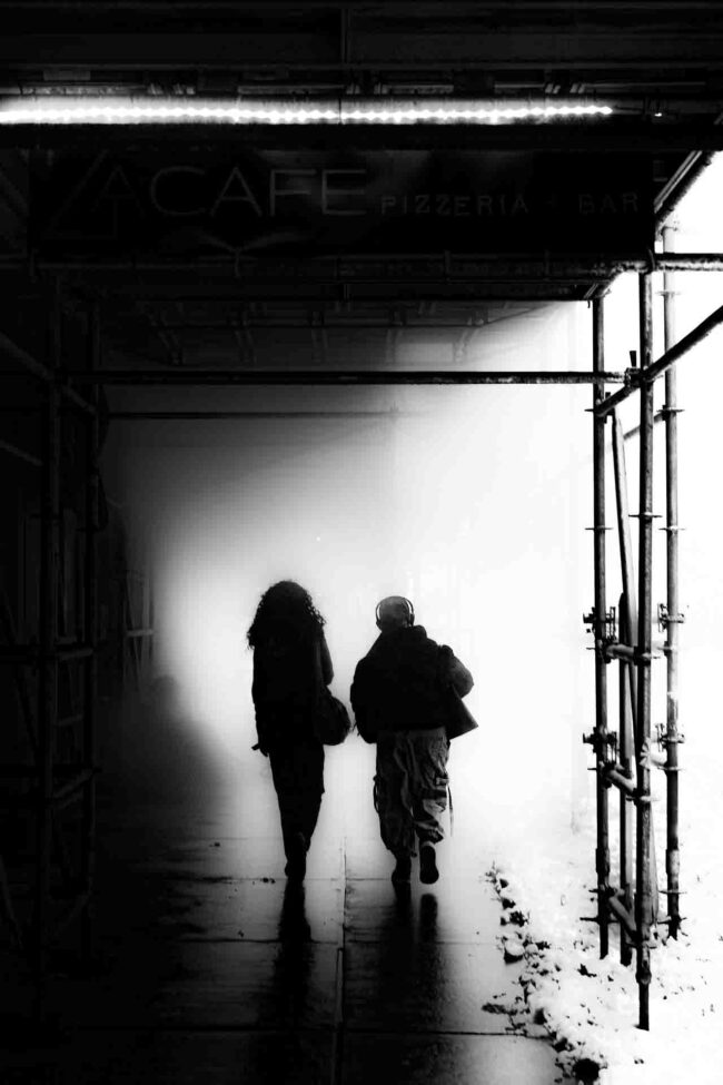 two females in black and white walking under a covered sidewalk walking towards a thick haze.