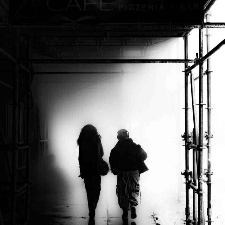 two females in black and white walking under a covered sidewalk walking towards a thick haze.