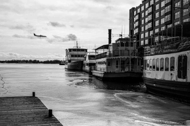 a black and white image of a semi-frozen lake with boats docked to its right and a plane landing to its left.