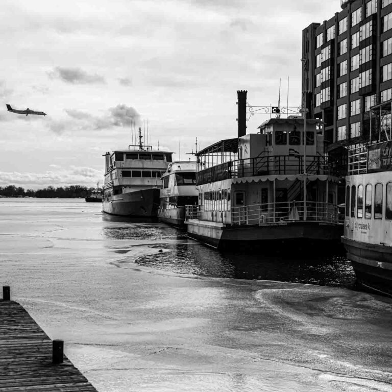 a black and white image of a semi-frozen lake with boats docked to its right and a plane landing to its left.